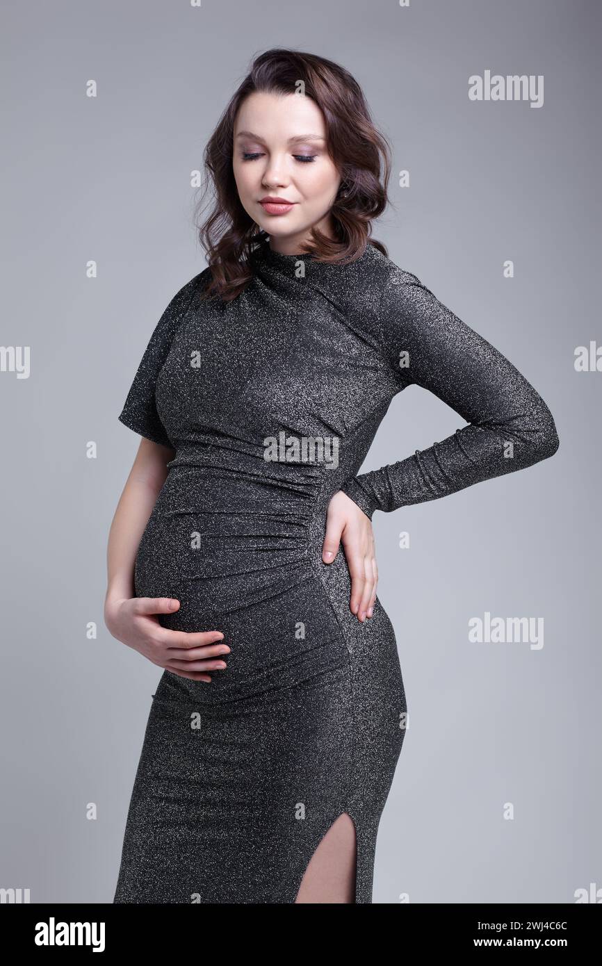 Ritratto di una giovane donna incinta in abito di paillettes grigio con le mani vicino alla pancia incinta. Foto Stock