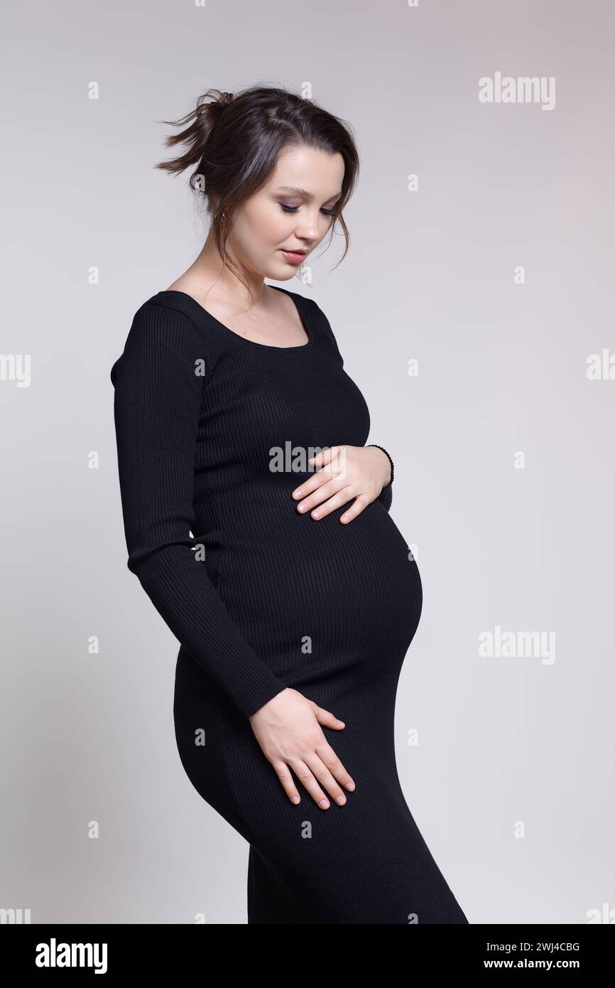 Ritratto di una giovane donna incinta in abito nero con le mani vicino alla pancia incinta. Foto Stock