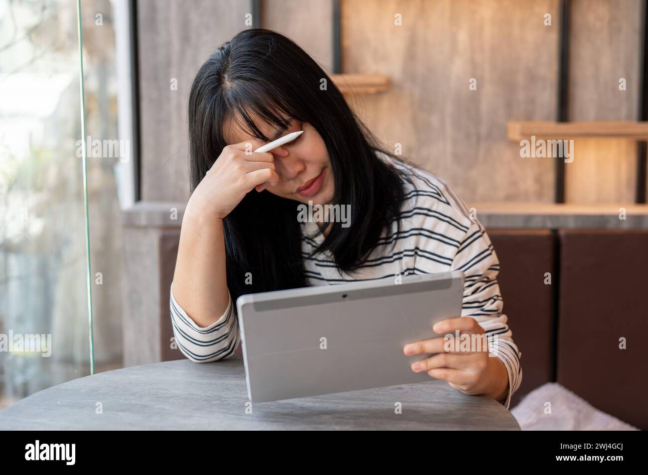 Una donna asiatica seria e delusa guarda lo schermo del suo tablet con un volto serio, contempla e risolve il problema lavorando da remoto Foto Stock