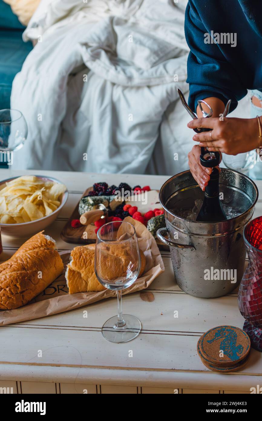 donna in camicia blu che apre una bottiglia di vino freddo nel secchio dello champagne sul tavolino da caffè con spuntini Foto Stock