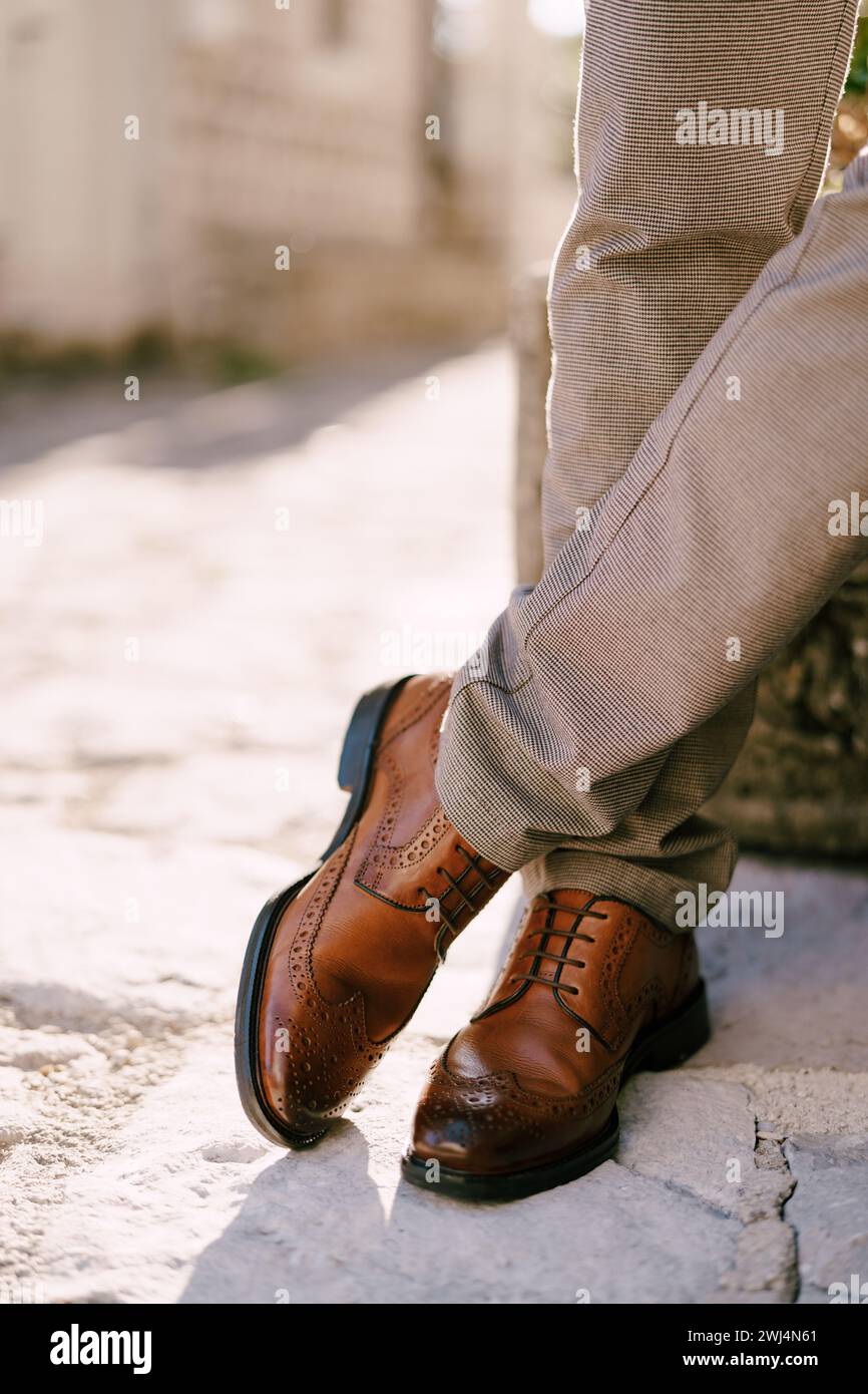 L'uomo con scarpe marroni si trova sulle pietre di pavimentazione con gambe incrociate. Ritagliato. Senza volto Foto Stock