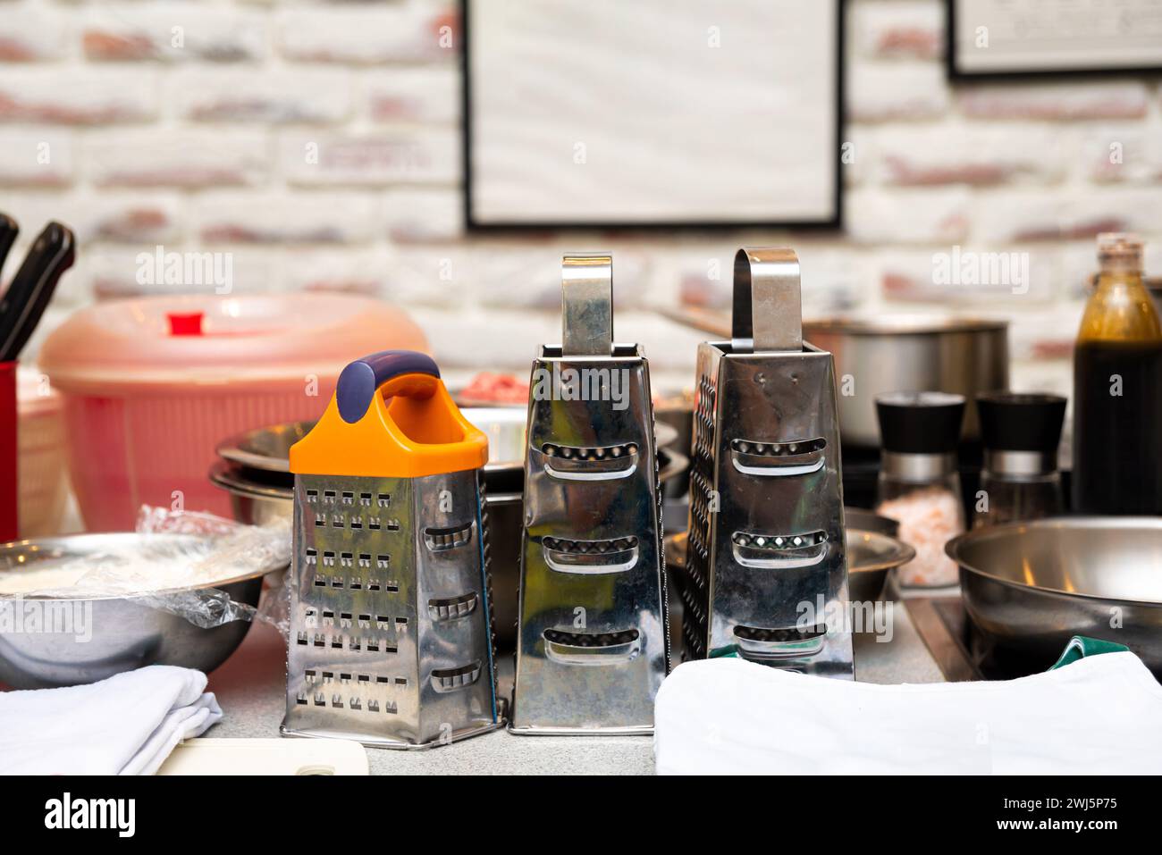 Un'affollata area di lavoro professionale con grattugie in acciaio inossidabile e vari utensili da cucina, con una parete in mattoni sullo sfondo. Foto Stock