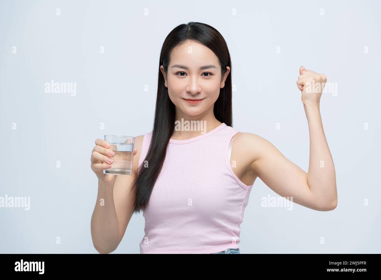 Sorridente giovane donna asiatica che regge un bicchiere di acqua pulita con il braccio sollevato su bianco Foto Stock