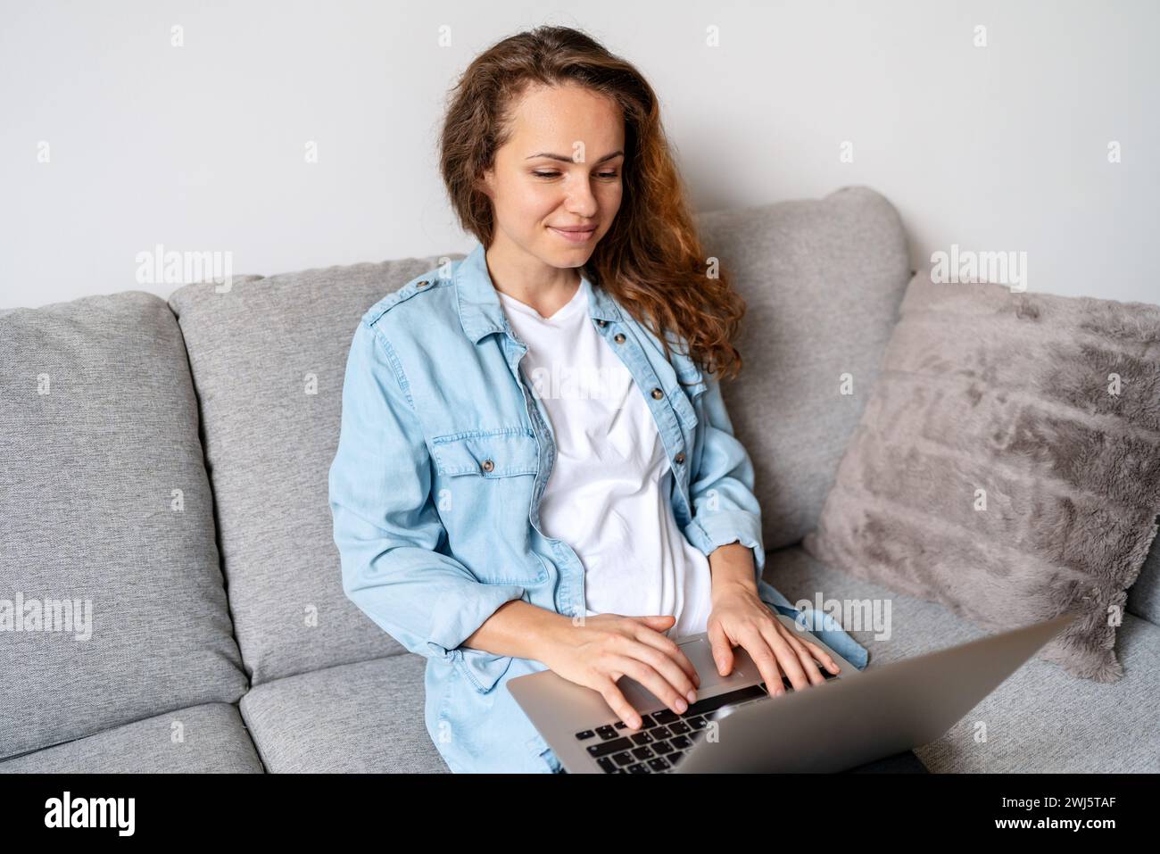 Donna bruna in camicia in denim seduta sul divano di casa e con un computer portatile. Shopping online. Lavoro da casa. Foto Stock