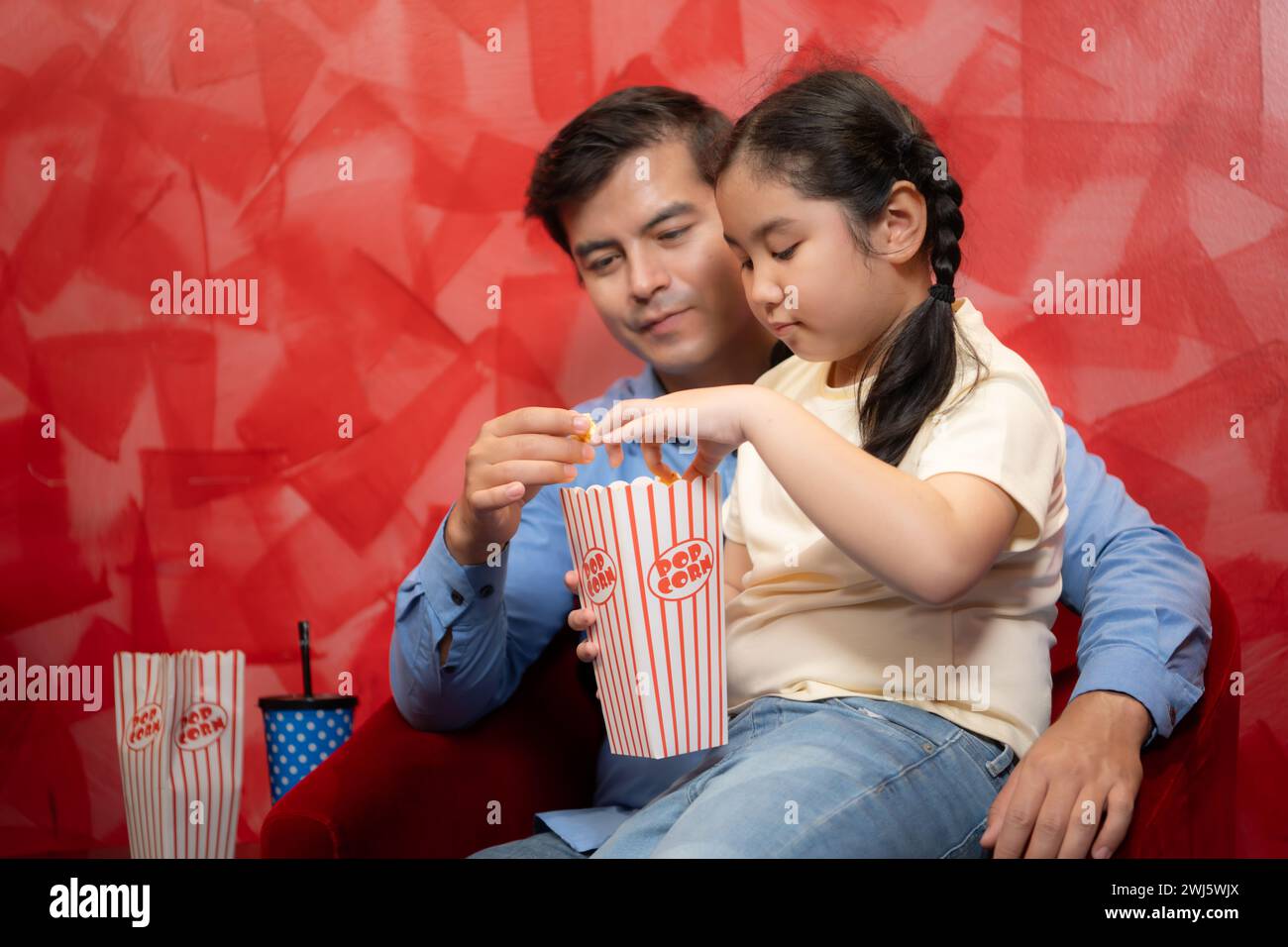 Padre e figlia mangiano popcorn e aspettano di guardare un film su sfondo rosso. Concetto di intrattenimento per famiglie. Foto Stock