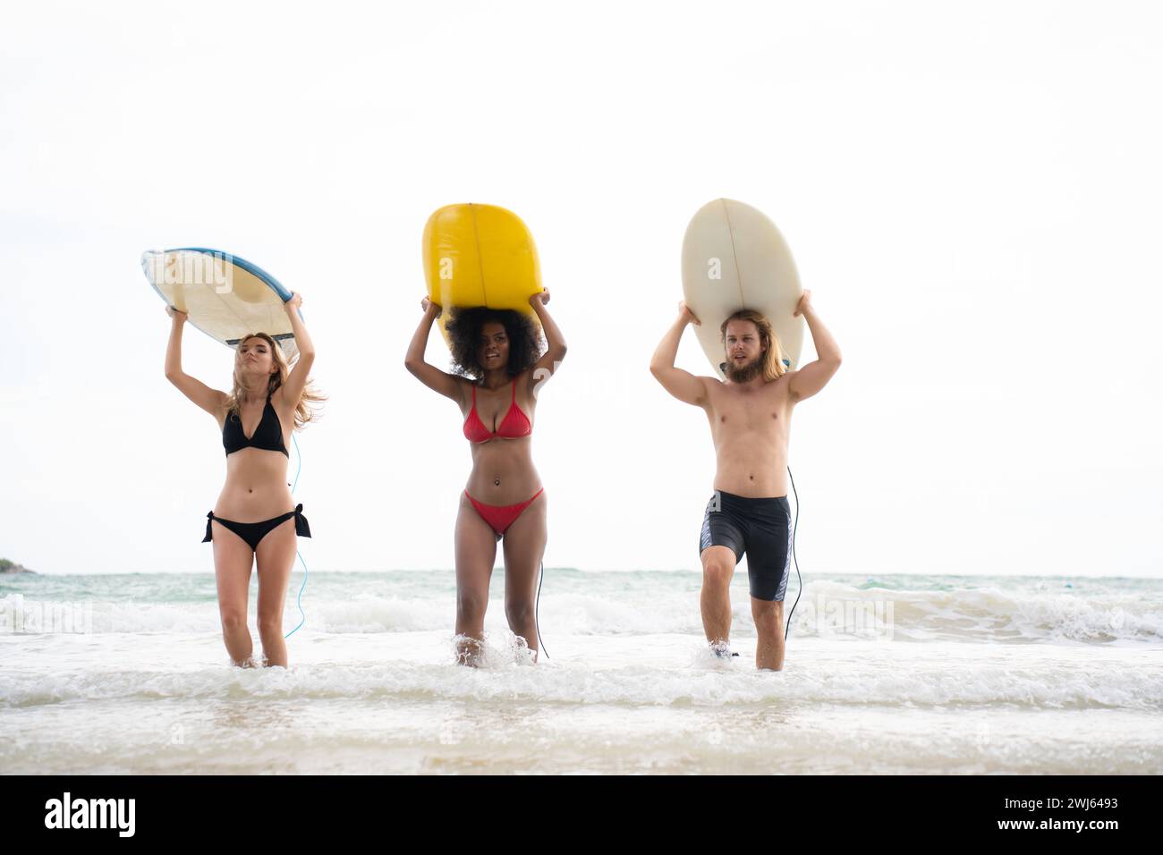 Vista posteriore di due donne e di un giovane che tengono le tavole da surf sulla testa e camminano verso il mare per fare surf Foto Stock