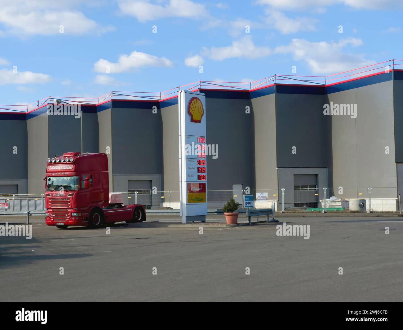 Amburgo, Germania - 26 febbraio 2022: Vista del camion Scania R500 parcheggiato vicino alla stazione di servizio Shell con i prezzi indicati e grande edificio di distribuzione del trasporto in magazzino sullo sfondo Foto Stock