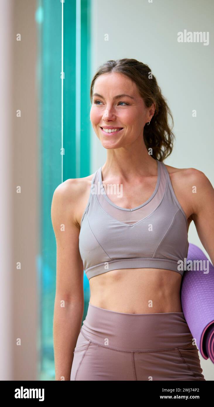 Ritratto di una donna che indossa un abbigliamento da palestra presso la palestra o lo studio di yoga tenendo in mano il tappetino per esercizi Foto Stock
