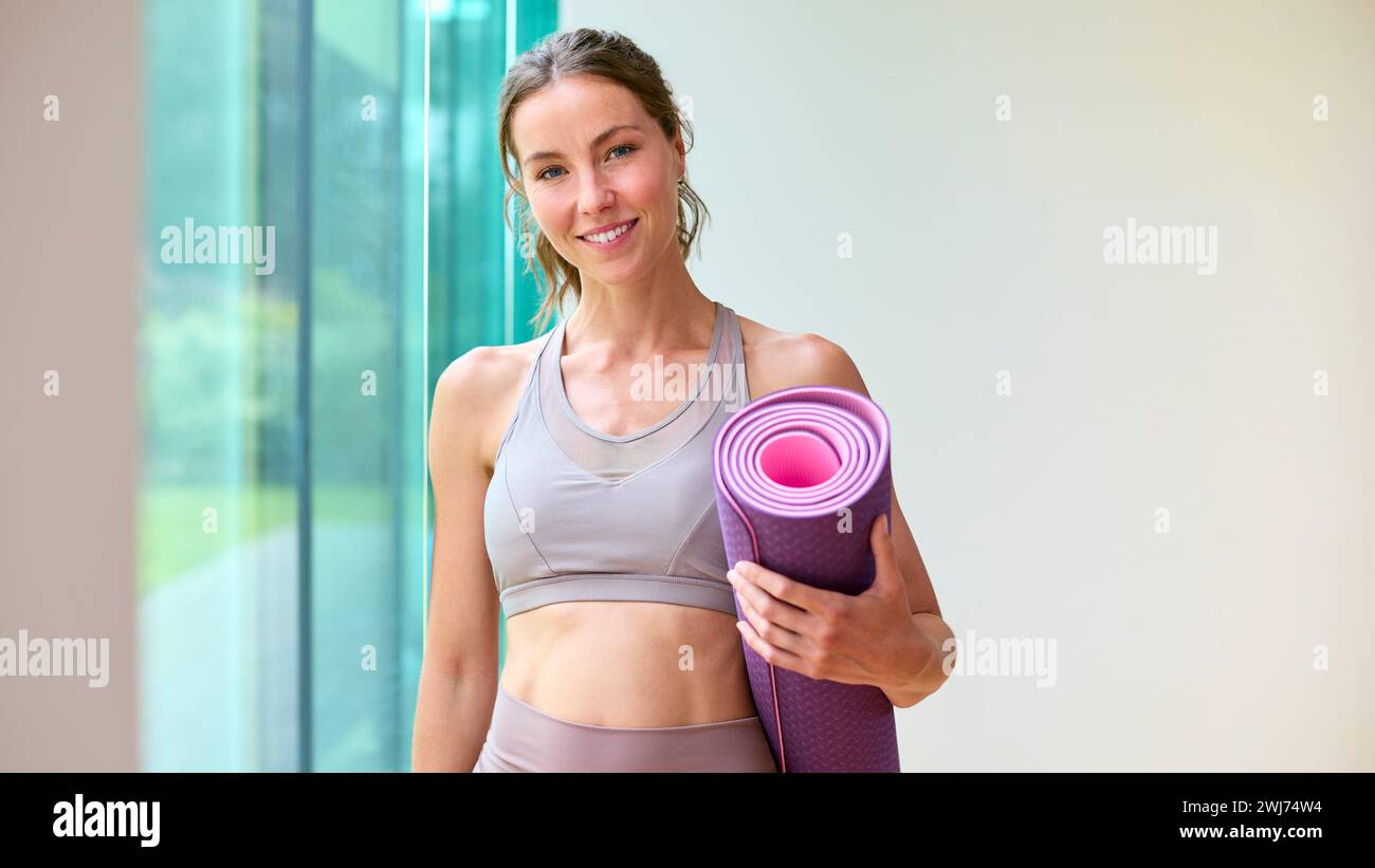 Ritratto di una donna che indossa un abbigliamento da palestra presso la palestra o lo studio di yoga tenendo in mano il tappetino per esercizi Foto Stock