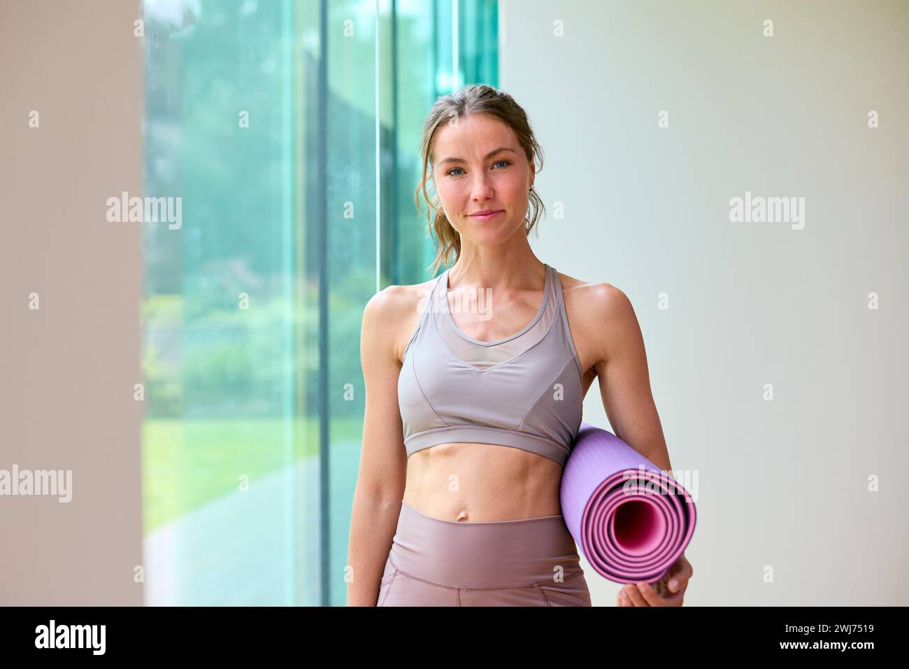 Ritratto di una donna che indossa un abbigliamento da palestra presso la palestra o lo studio di yoga tenendo in mano il tappetino per esercizi Foto Stock