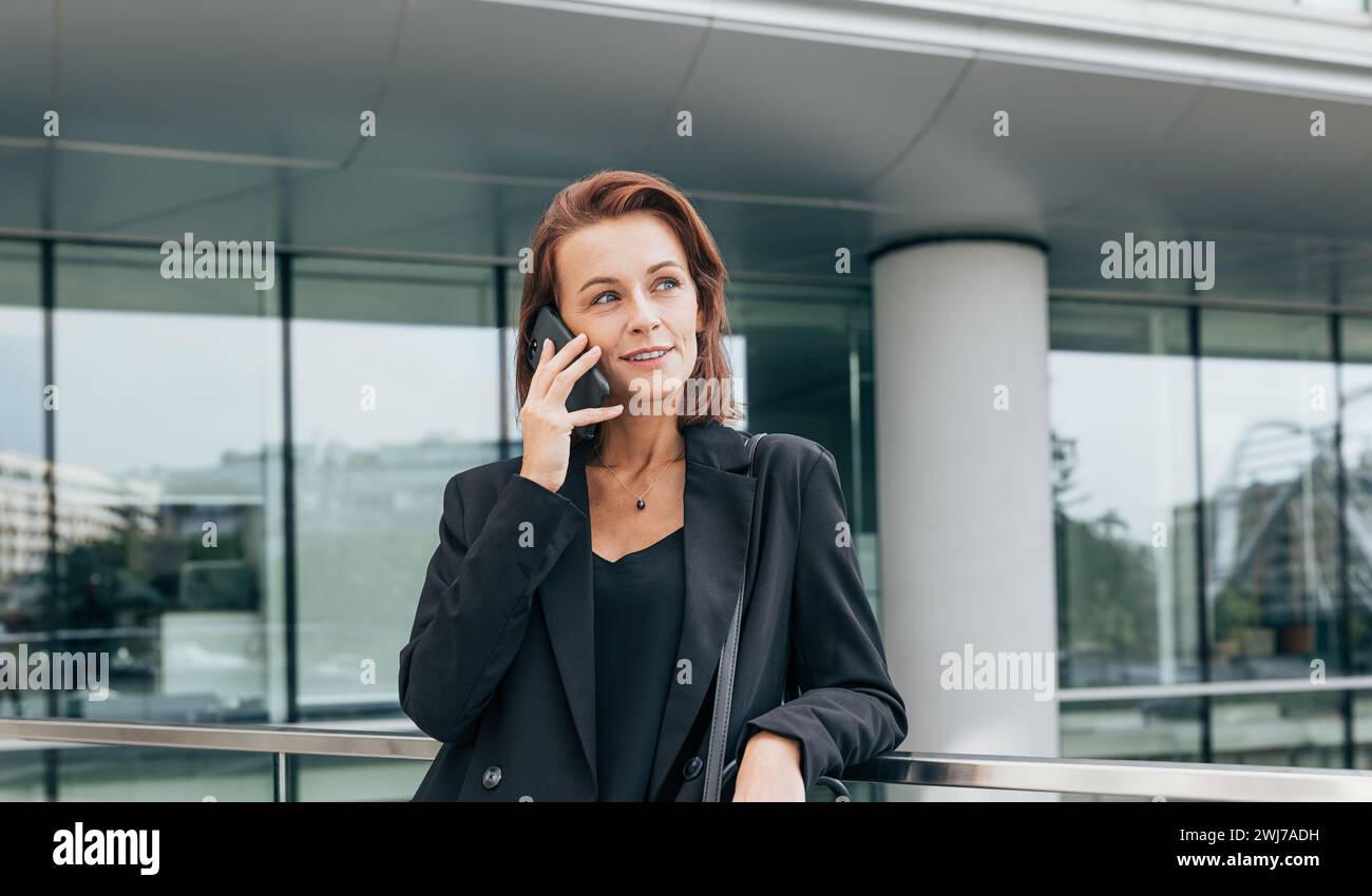 Donna di mezza età con capelli di zenzero che si distoglie mentre parla al telefono cellulare. Donna sicura di sé nell'abbigliamento formale in piedi nell'edificio dell'ufficio. Foto Stock