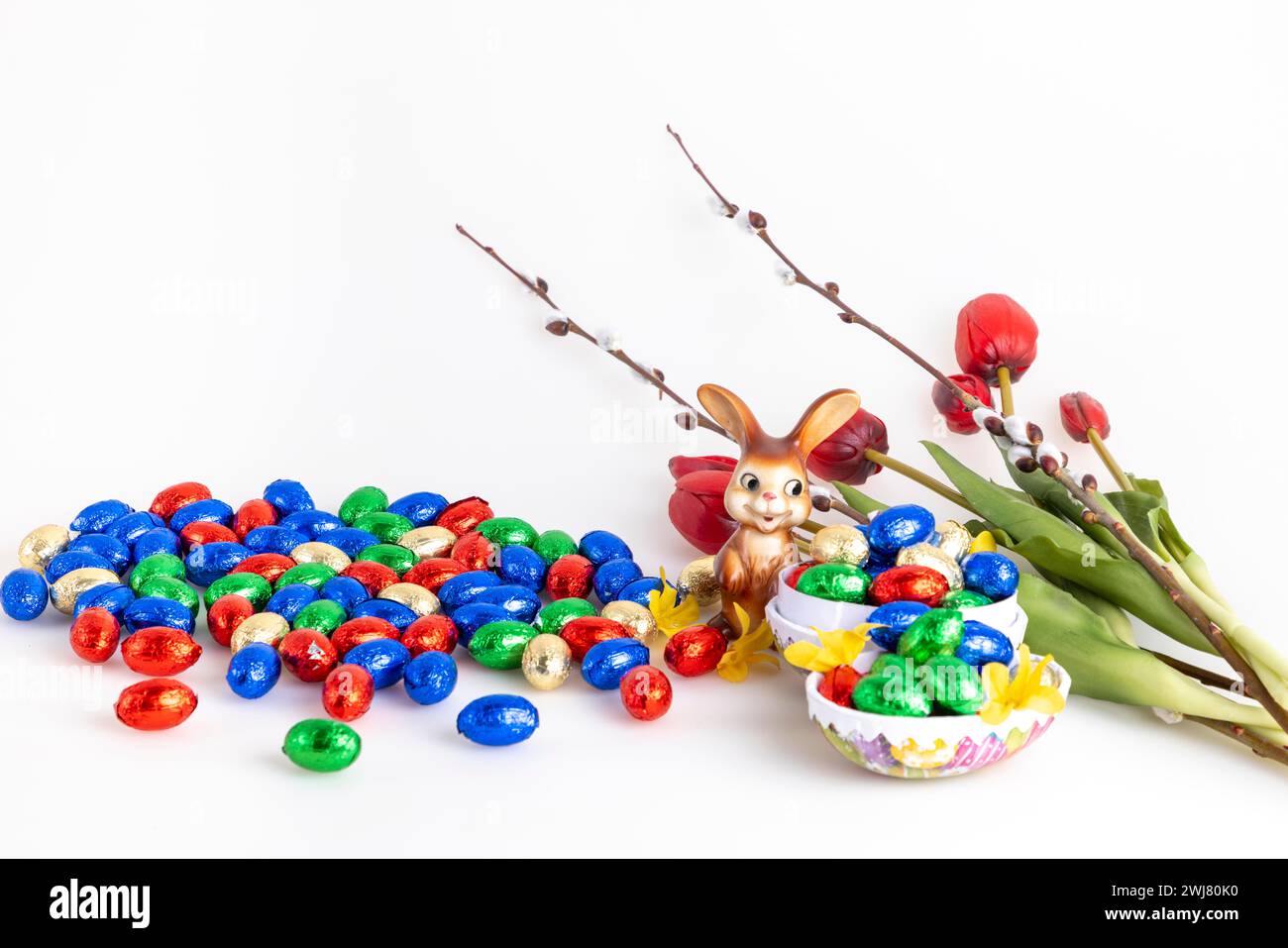 Una collezione di uova di cioccolato colorate e un coniglietto pasquale accanto a tulipani e gatti di palma, sfondo bianco Foto Stock