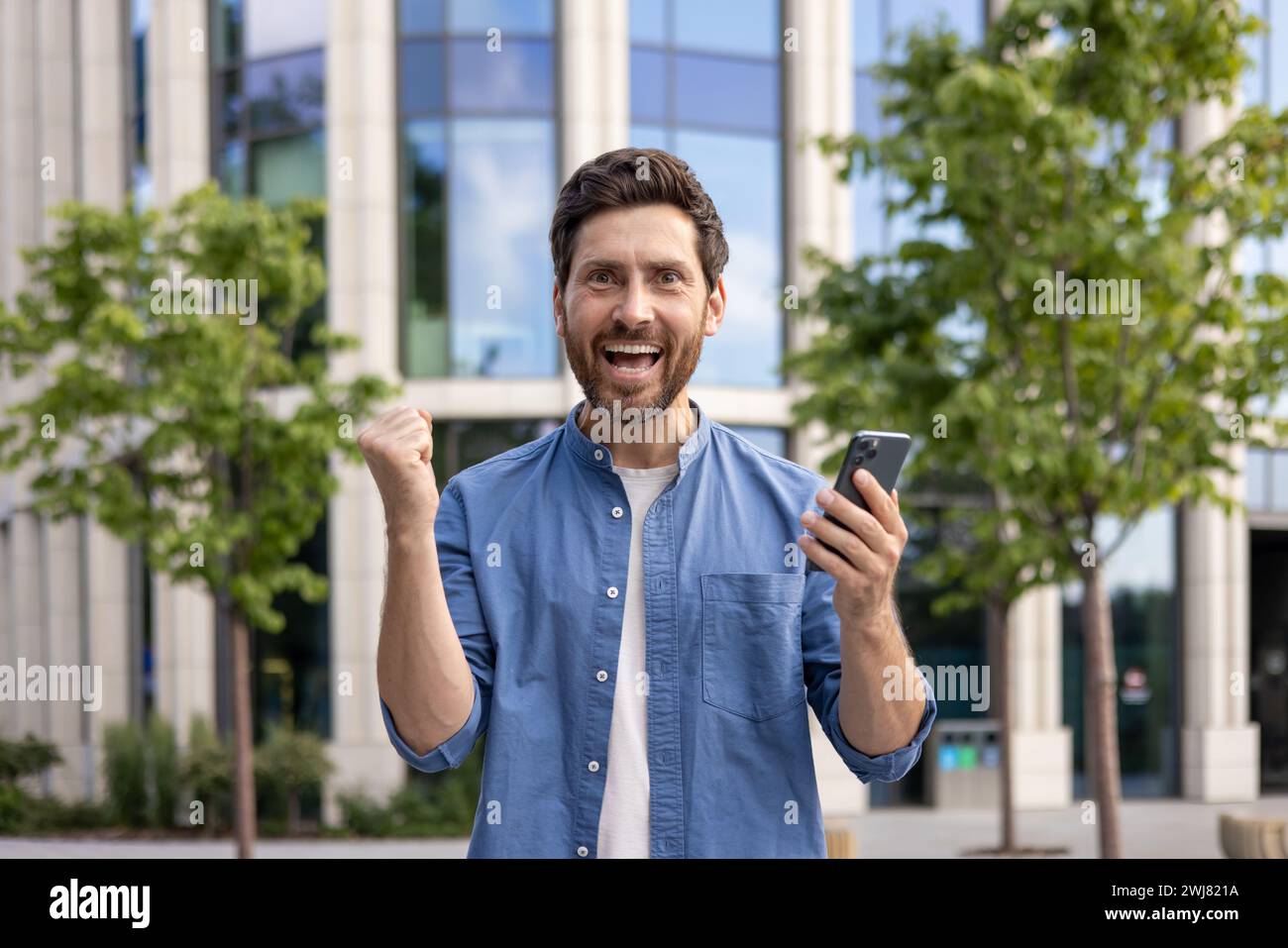 Ritratto ravvicinato di un giovane che si rallegra alla telecamera, mostra un segno di vittoria con le mani, tiene in mano un cellulare. Foto Stock