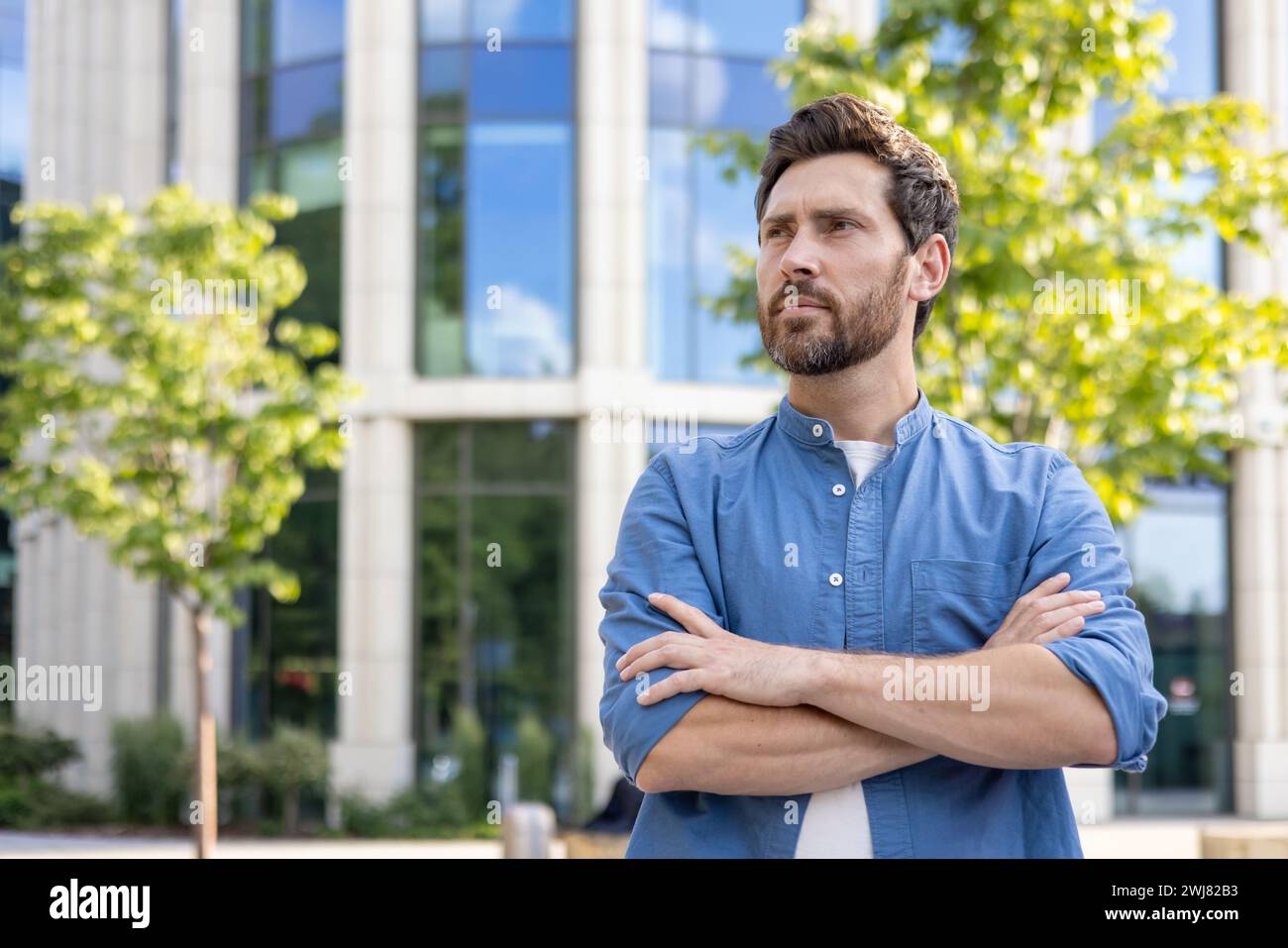 Uomo con barba matura in camicia casual con braccia ripiegate, dall'aspetto premuroso fuori da un ufficio. Foto Stock