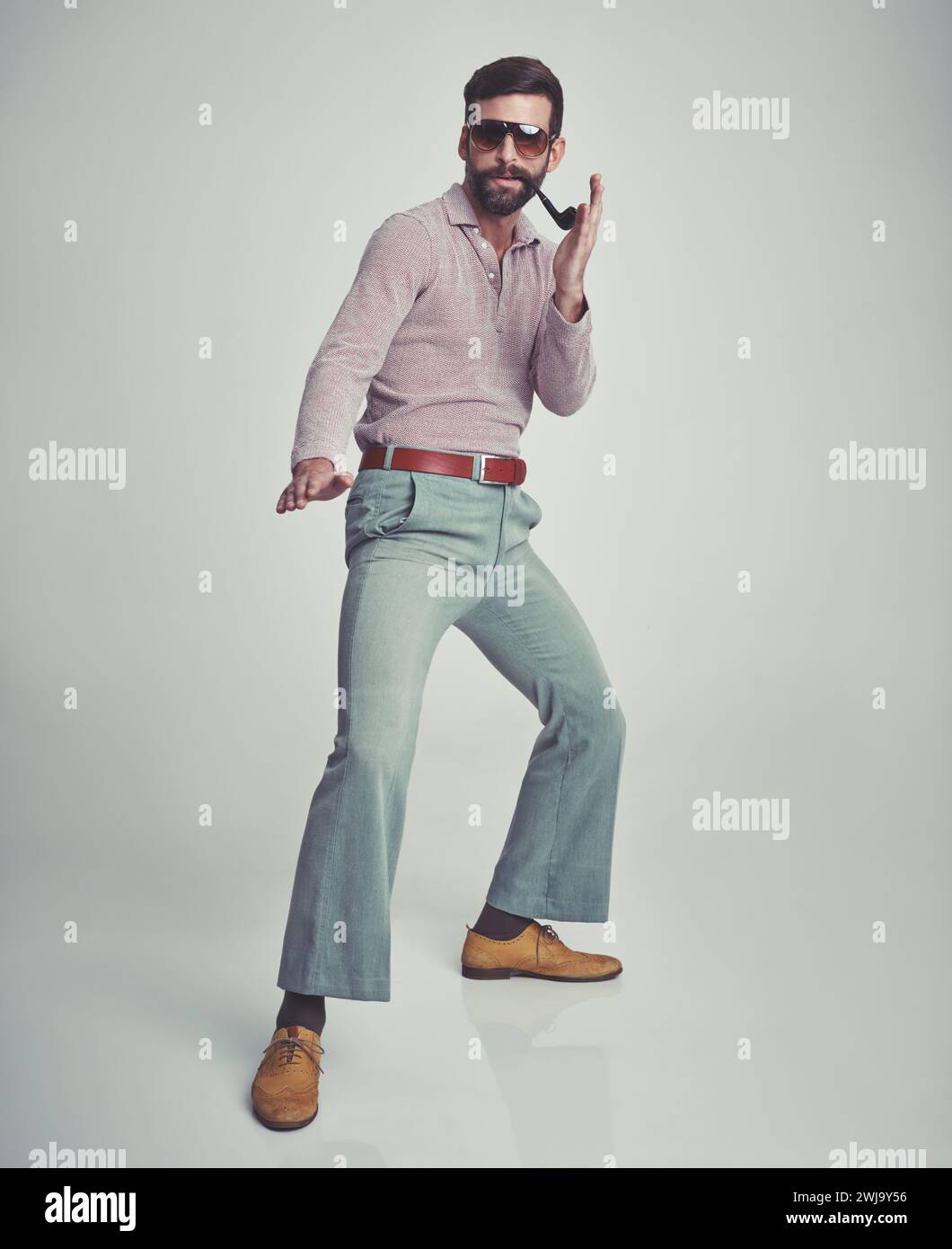 anni '70, moda e ritratto dell'uomo con un'estetica retrò o vintage su sfondo grigio dello studio. Fumare, pipa e persona ballano con fiducia nel funky Foto Stock