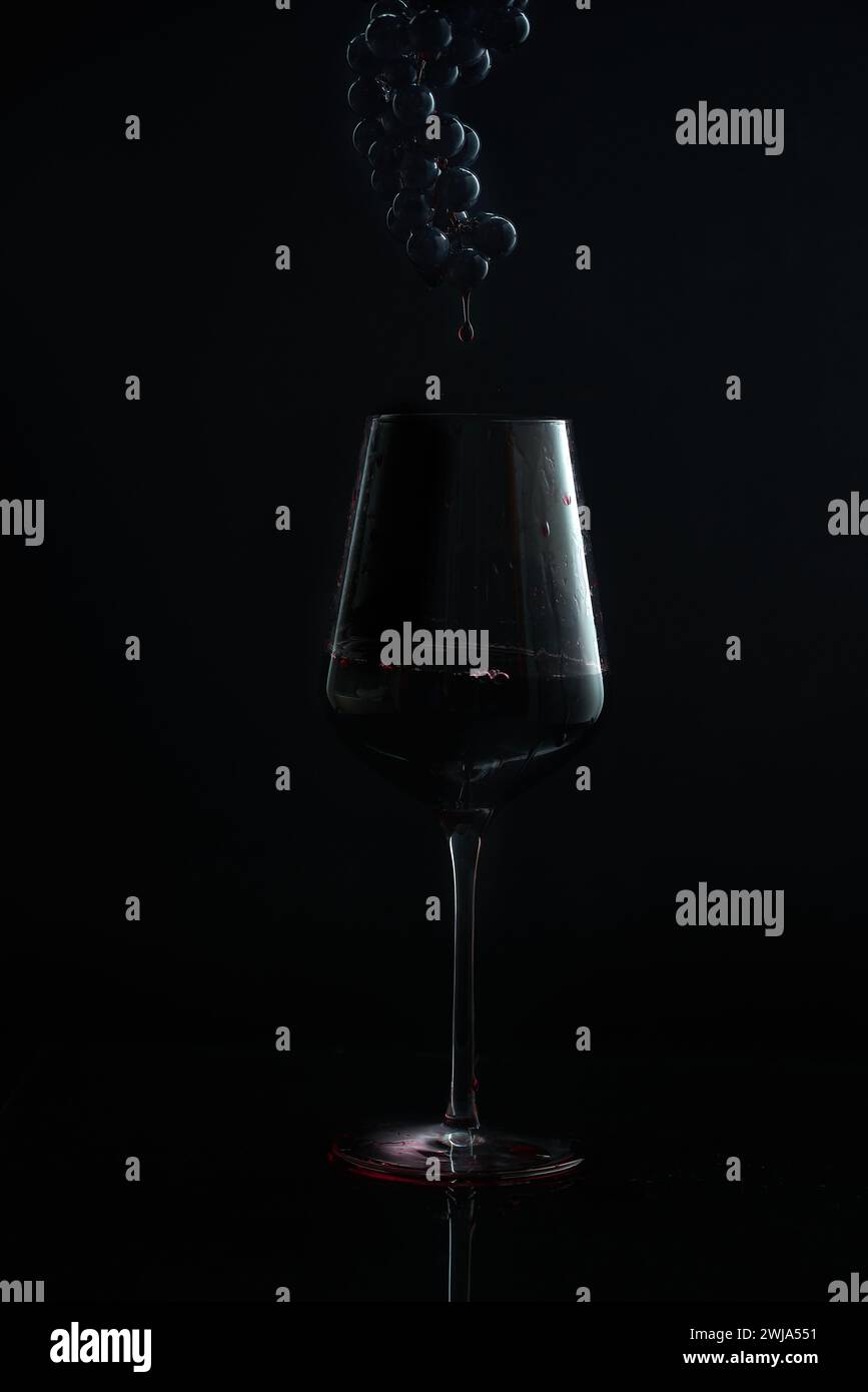 Uno scatto moody che cattura un triclo di vino che scende in un bicchiere con un grappolo d'uva scuro che si libra sopra su uno sfondo nero Foto Stock