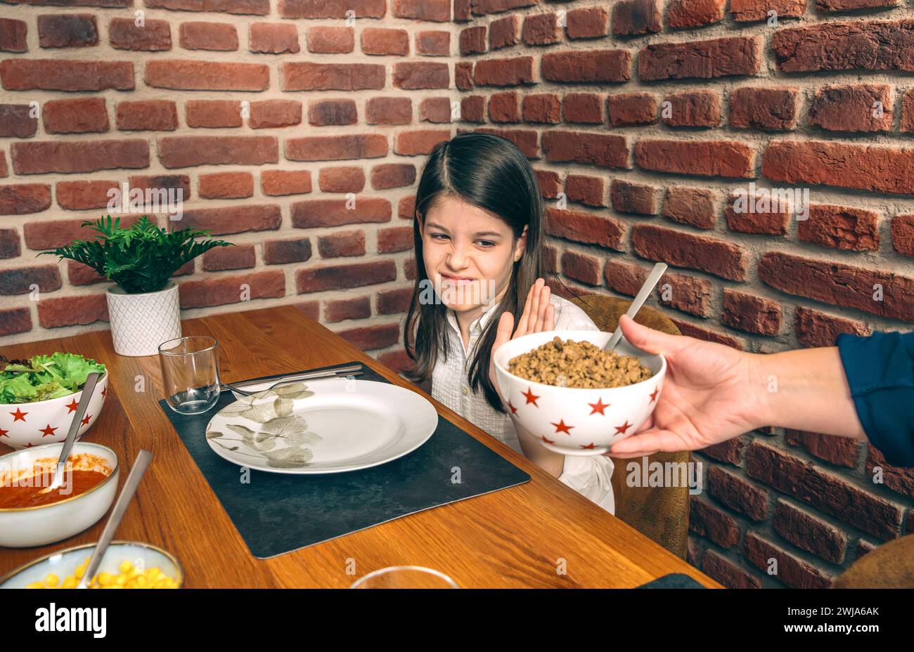 Una giovane ragazza guarda scetticamente una ciotola di cibo che le viene offerta al tavolo da pranzo di famiglia su uno sfondo di un muro di mattoni Foto Stock