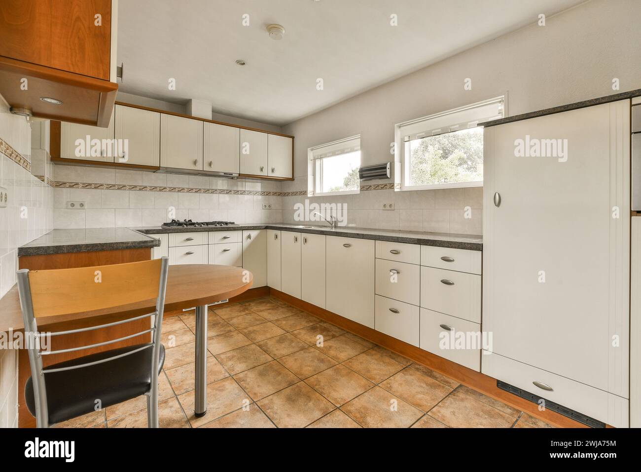 Una ben illuminata e spaziosa cucina con armadi in legno bianco, pavimento piastrellato marrone e una piccola area da pranzo con tavolo e sedia. Foto Stock