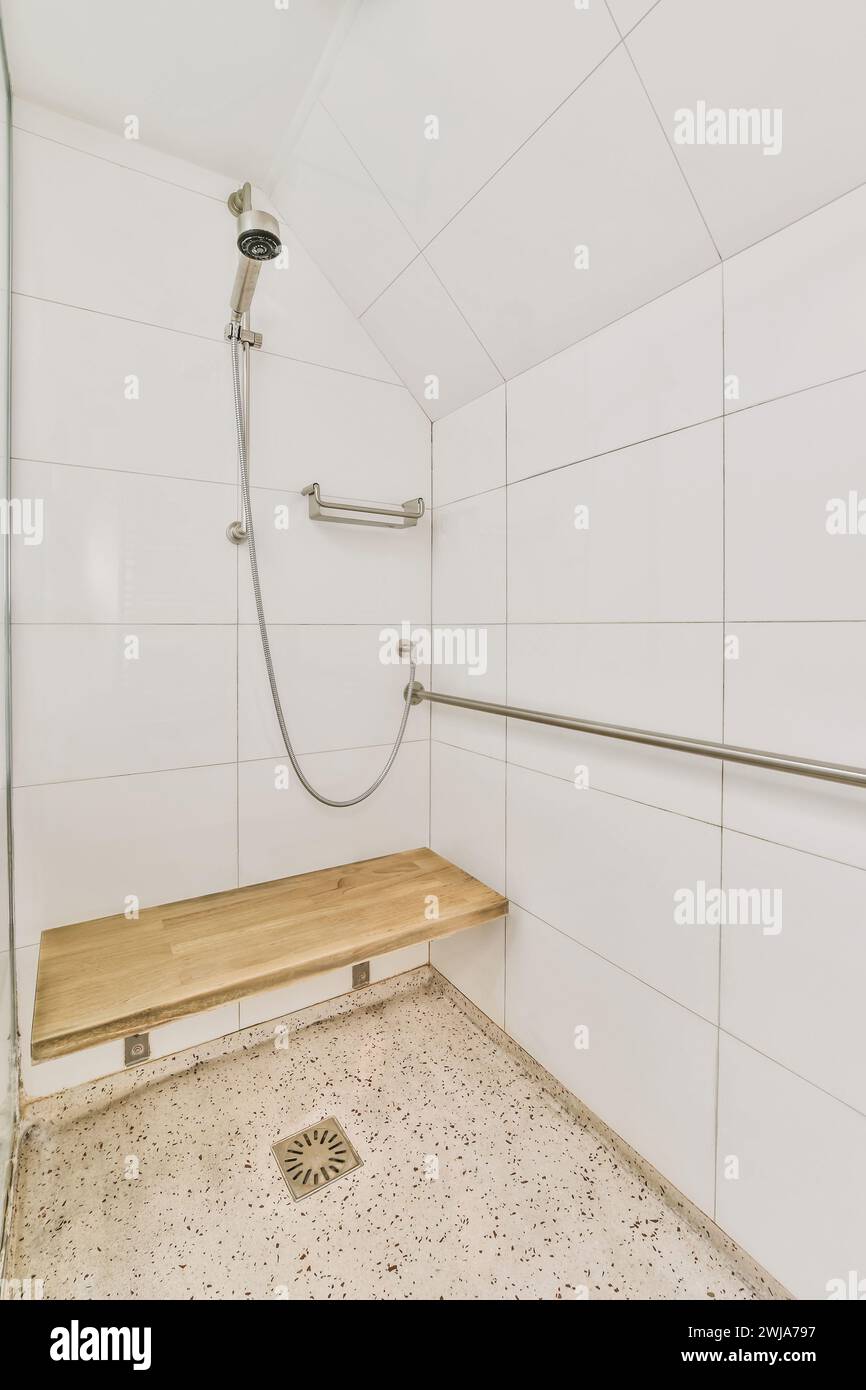 Un moderno spazio doccia con panca in legno e piastrelle in ceramica bianca, con soffione doccia, tubo flessibile e barre di appoggio per la sicurezza. Foto Stock