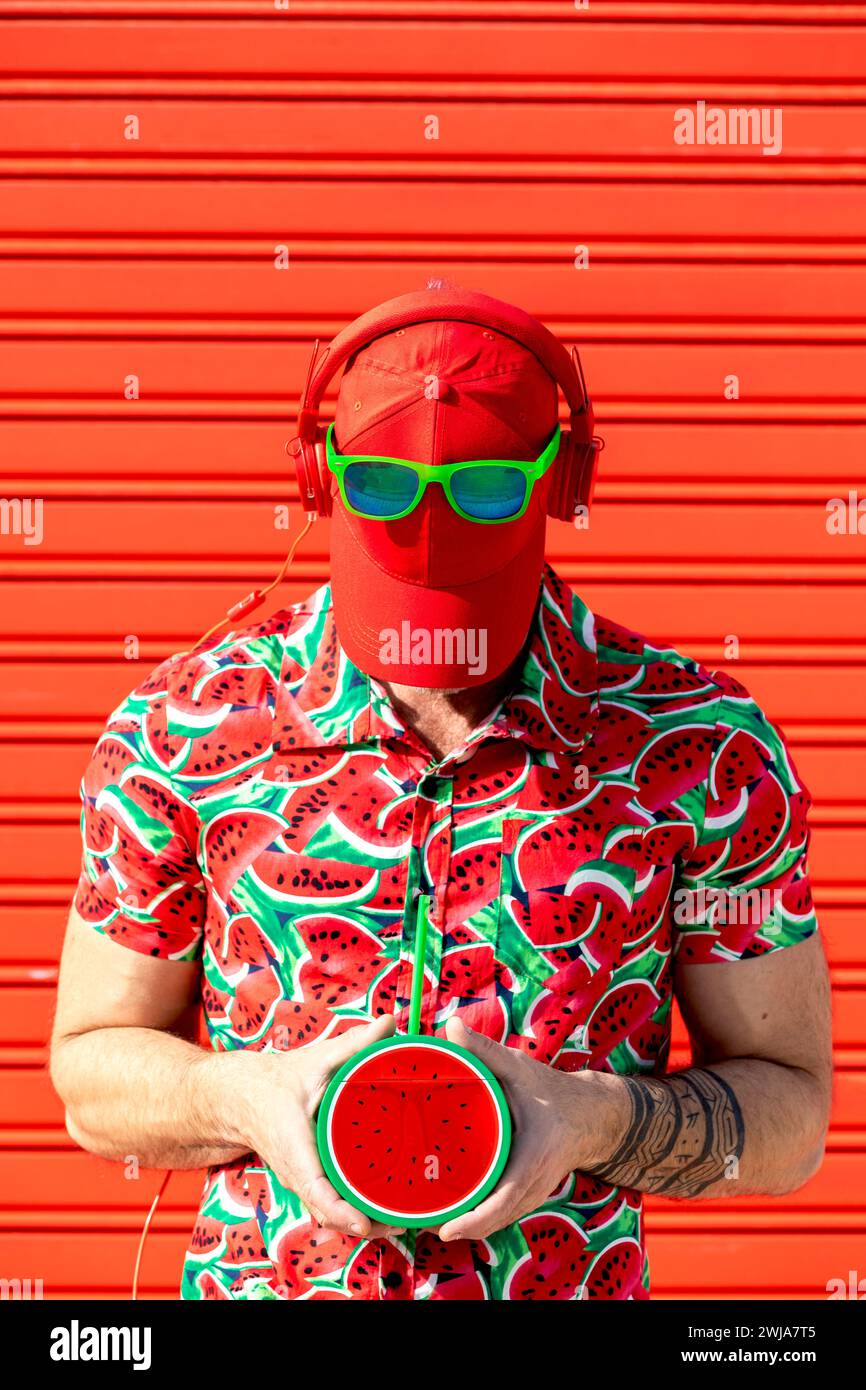 Uomo Street style irriconoscibile con cappello che copre il viso e cuffie da sole in posizione mentre si sta in piedi nella camicia stampata con fette di anguria Foto Stock