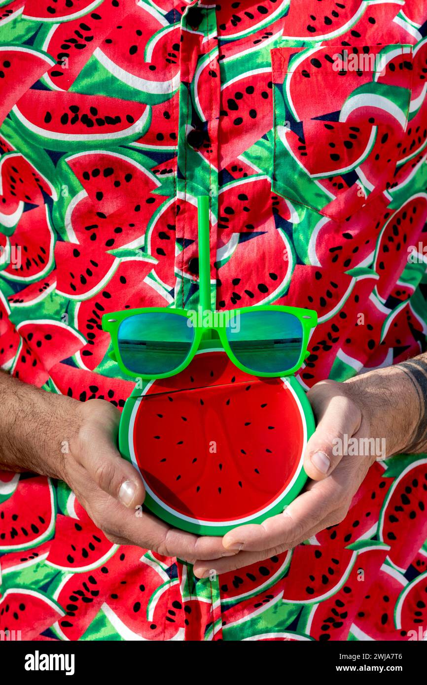 Uomo anonimo in stile Street mentre indossa una camicia stampata alla moda con angurie affettate alla luce del sole che trasportano succo di finta con paglia e occhiali da sole Foto Stock