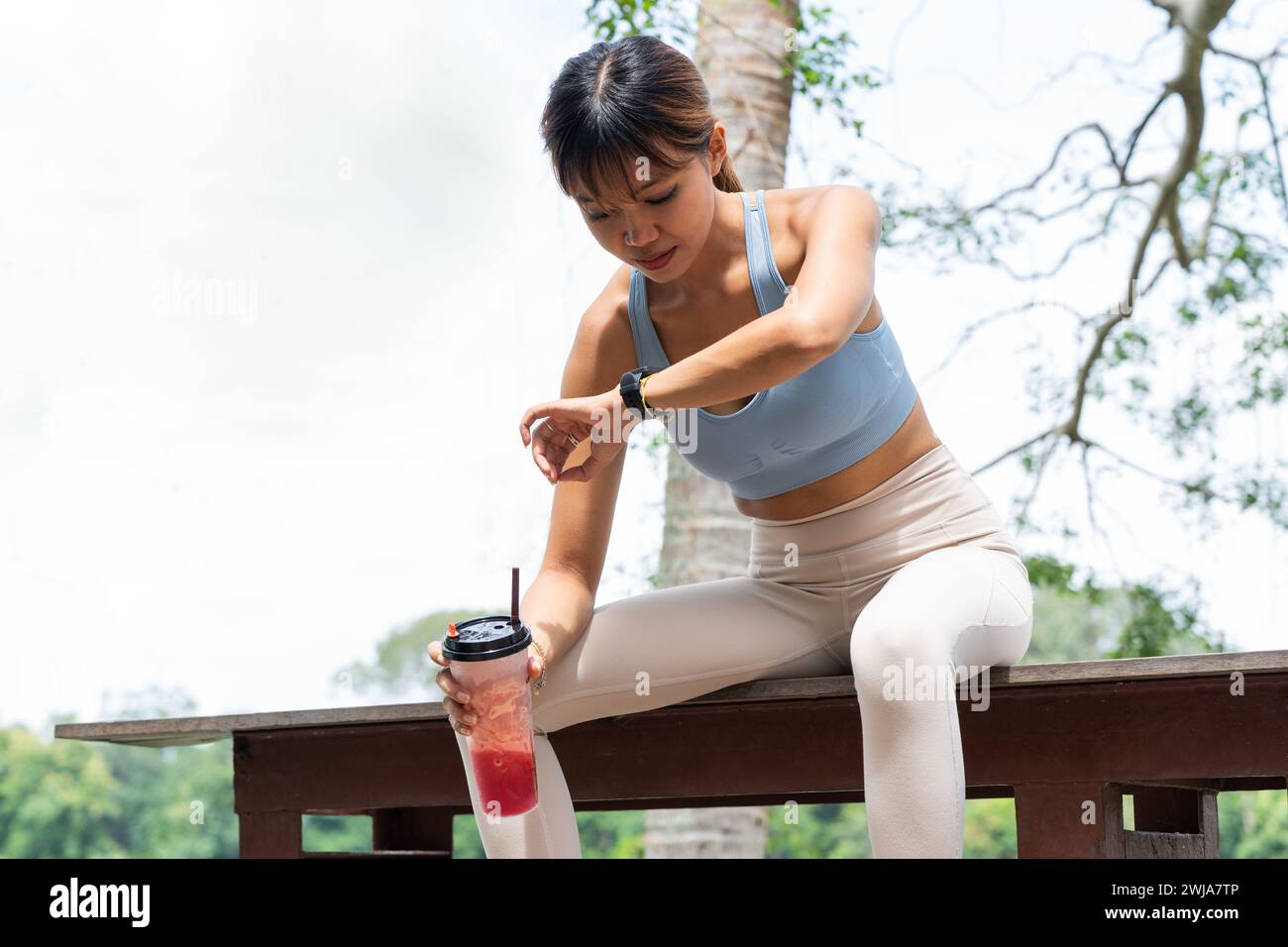 Contenta la giovane donna bruna in abbigliamento sportivo seduta sulla panchina nel parco mentre beve dalla tazza di plastica e usa lo smartwatch per controllare l'ora del sole Foto Stock