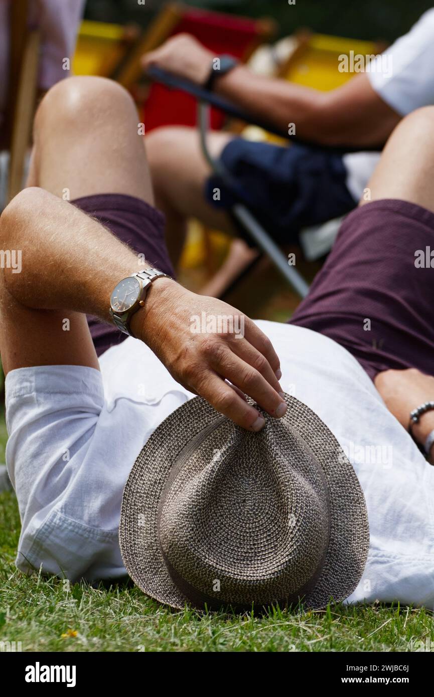 Uomo con la mano su Un trilby di paglia che si rilassa sull'erba durante Un concerto di musica dal vivo in estate, Regno Unito Foto Stock