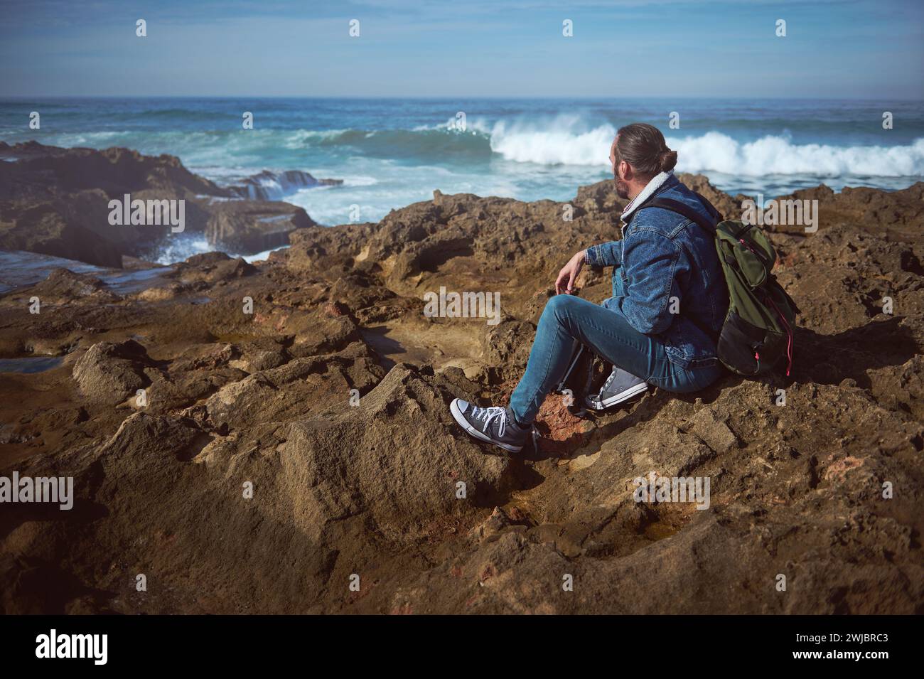 Bel turista che si rilassa su una scogliera rocciosa, affacciato sullo splendido paesaggio oceanico. Giovane viaggiatore avventuriero seduto da solo, ammirando le terre Foto Stock