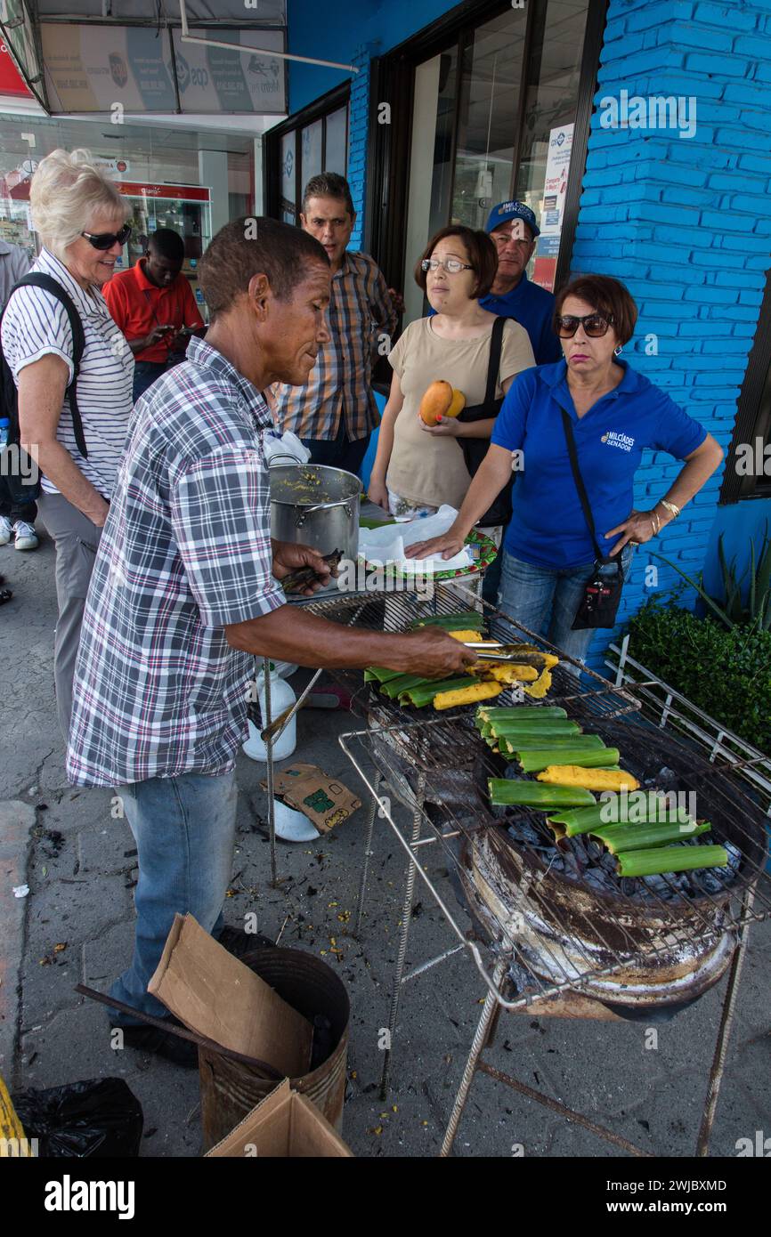 Un uomo che cucina tamales domenicani su una griglia aperta in strada al Bani Mango Expo di Bani, Repubblica Dominicana. Un bel tour biondo senior Foto Stock