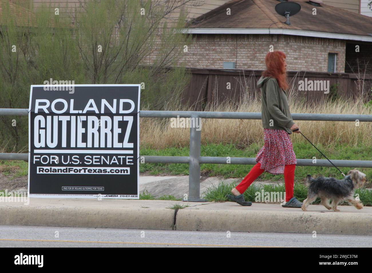 San Antonio, Stati Uniti. 14 febbraio 2024. Una donna che cammina con il suo cane passa accanto a un cartello della campagna di Roland Gutierrez lungo la Medina base Road a San Antonio, Texas, USA, il 14 febbraio 2024. Roland Gutierrez è attualmente un membro del Senato del Texas e il suo distretto comprende Uvalde, Texas. Dopo la sparatoria di massa della scuola elementare Robb, ha introdotto quattro bollette di sicurezza per le armi. (Foto di Carlos Kosienski/Sipa USA) credito: SIPA USA/Alamy Live News Foto Stock