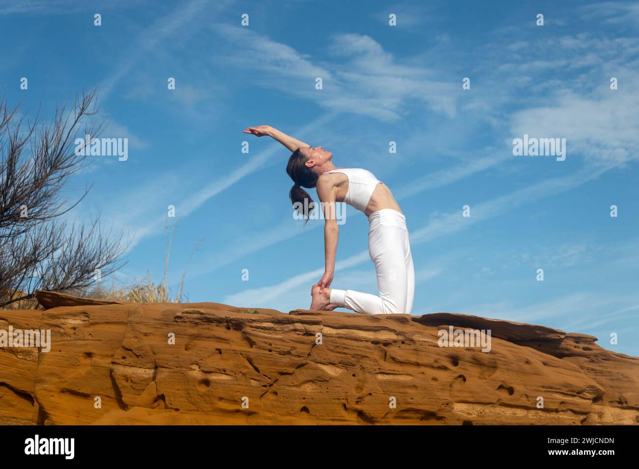 donna in forma che fa una posa di yoga a dorso di cammello su rocce rosse, sfondo blu del cielo Foto Stock