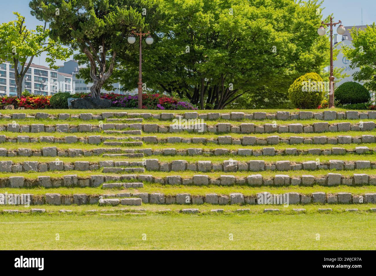 Posti a sedere a schiera fatti di massi davanti a un boschetto di alberi e fiori nell'anfiteatro del parco pubblico della Corea del Sud Foto Stock