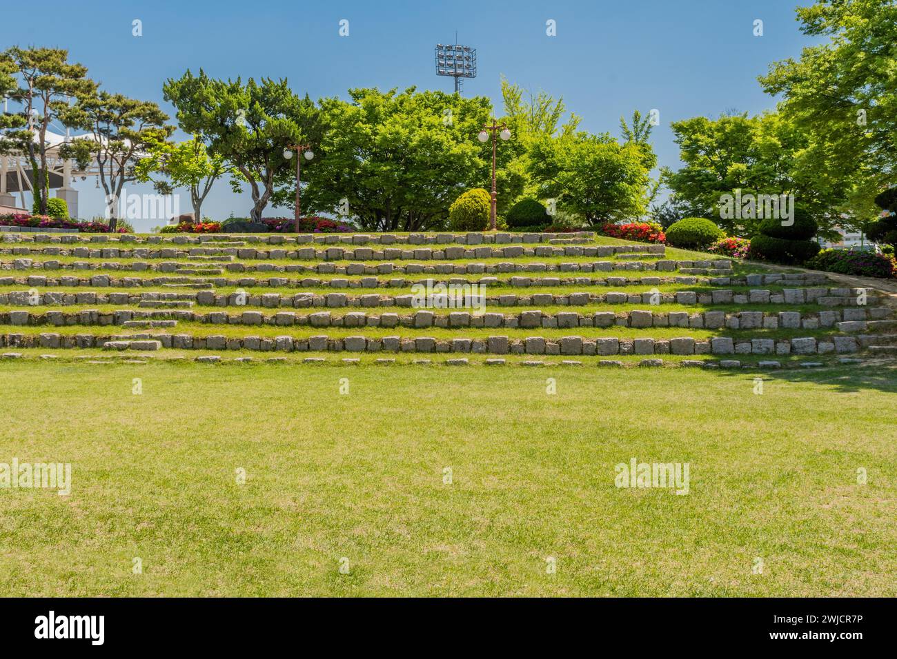 Posti a sedere a schiera fatti di massi davanti a un boschetto di alberi e fiori nell'anfiteatro del parco pubblico della Corea del Sud Foto Stock