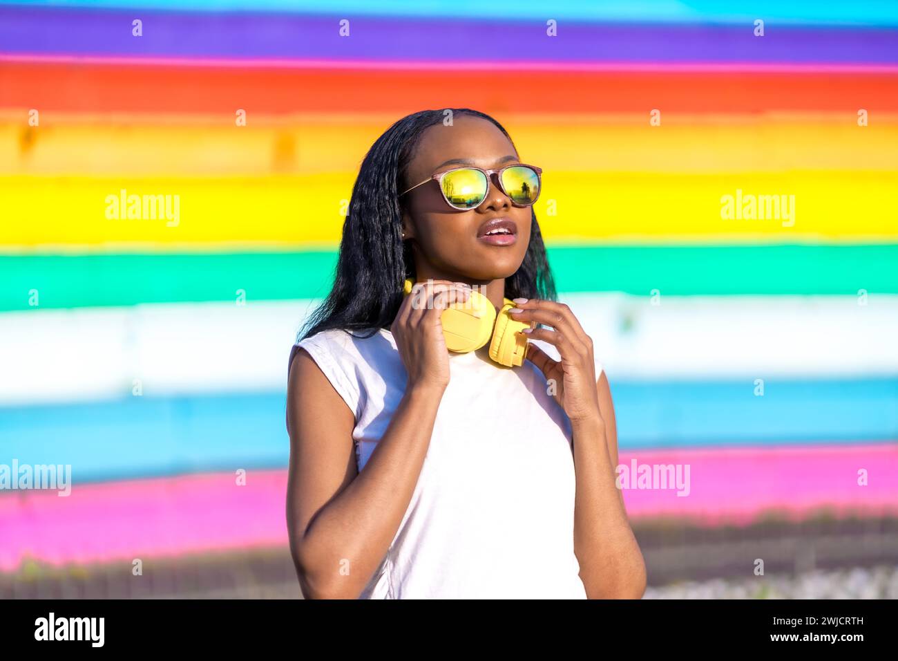 Elegante donna africana distratta che indossa occhiali da sole e cuffie in un parco urbano multicolore Foto Stock