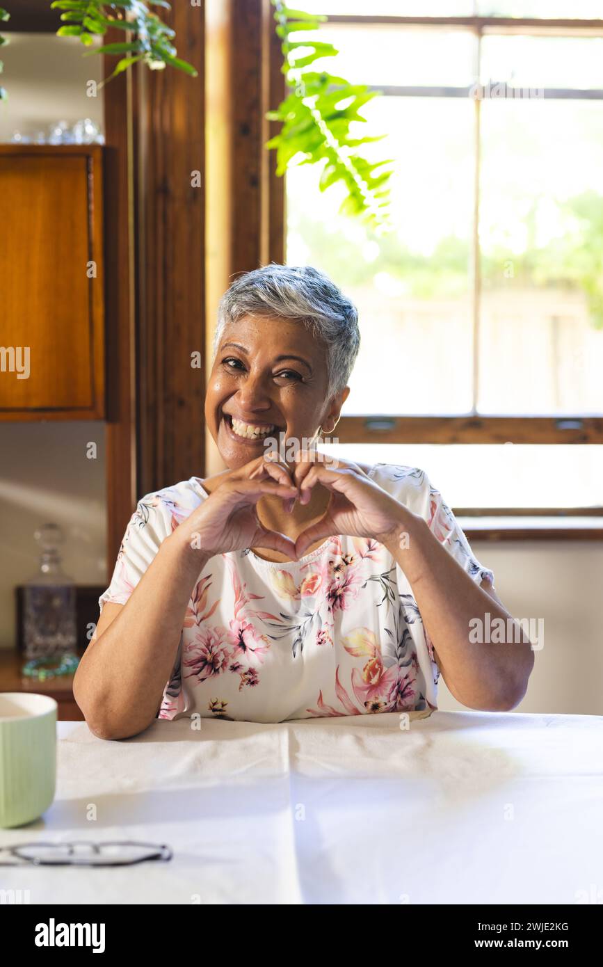 Una donna birazziale matura è allegra e crea una forma a cuore con le mani a casa durante una videochiamata Foto Stock