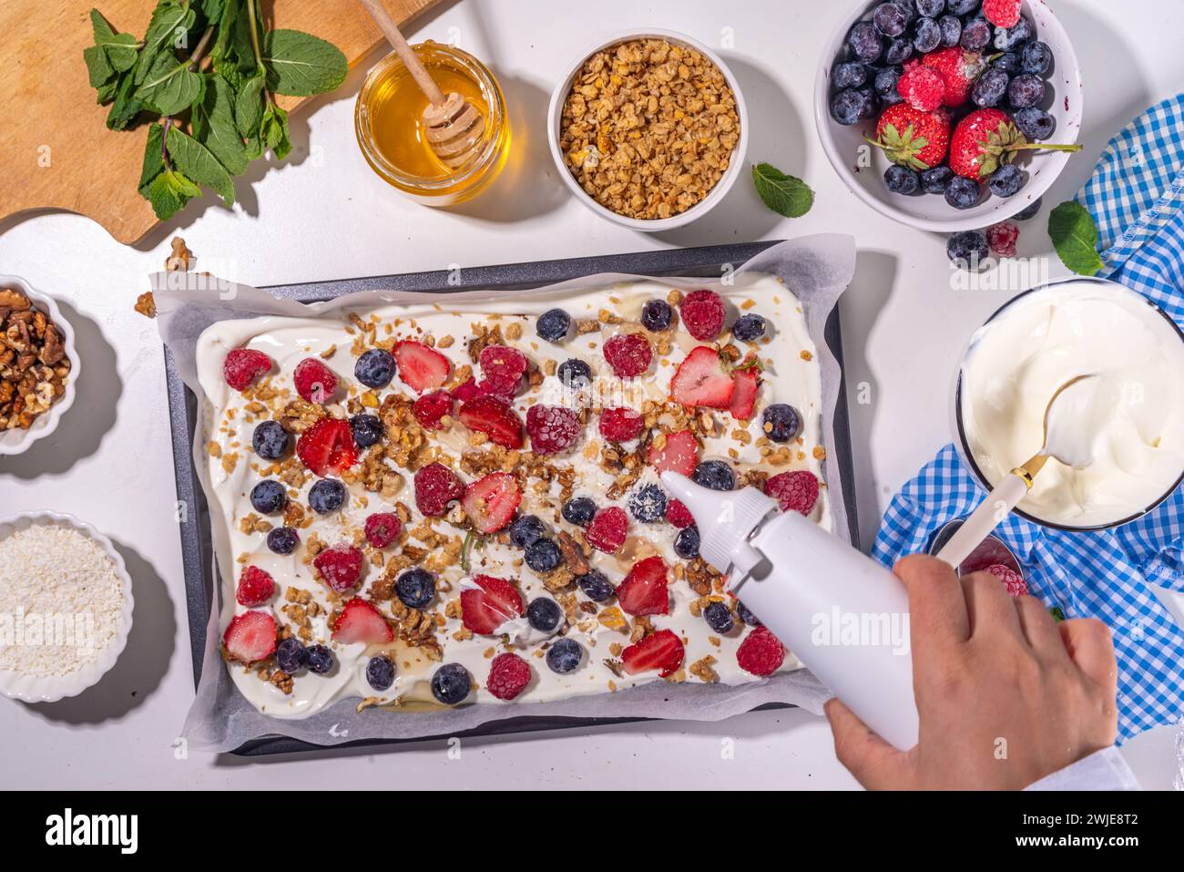 Preparazione della cottura con sfondo di corteccia di yogurt congelato con yogurt greco, bacche fresche, muesli, miele, salsa al cioccolato, con vassoio da forno deco. Vista dall'alto della donna Foto Stock