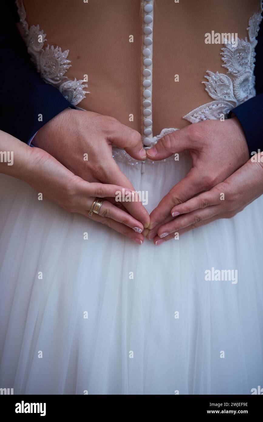 Un primo piano delle mani della sposa e dello sposo che formano il cuore sull'abito da sposa Foto Stock