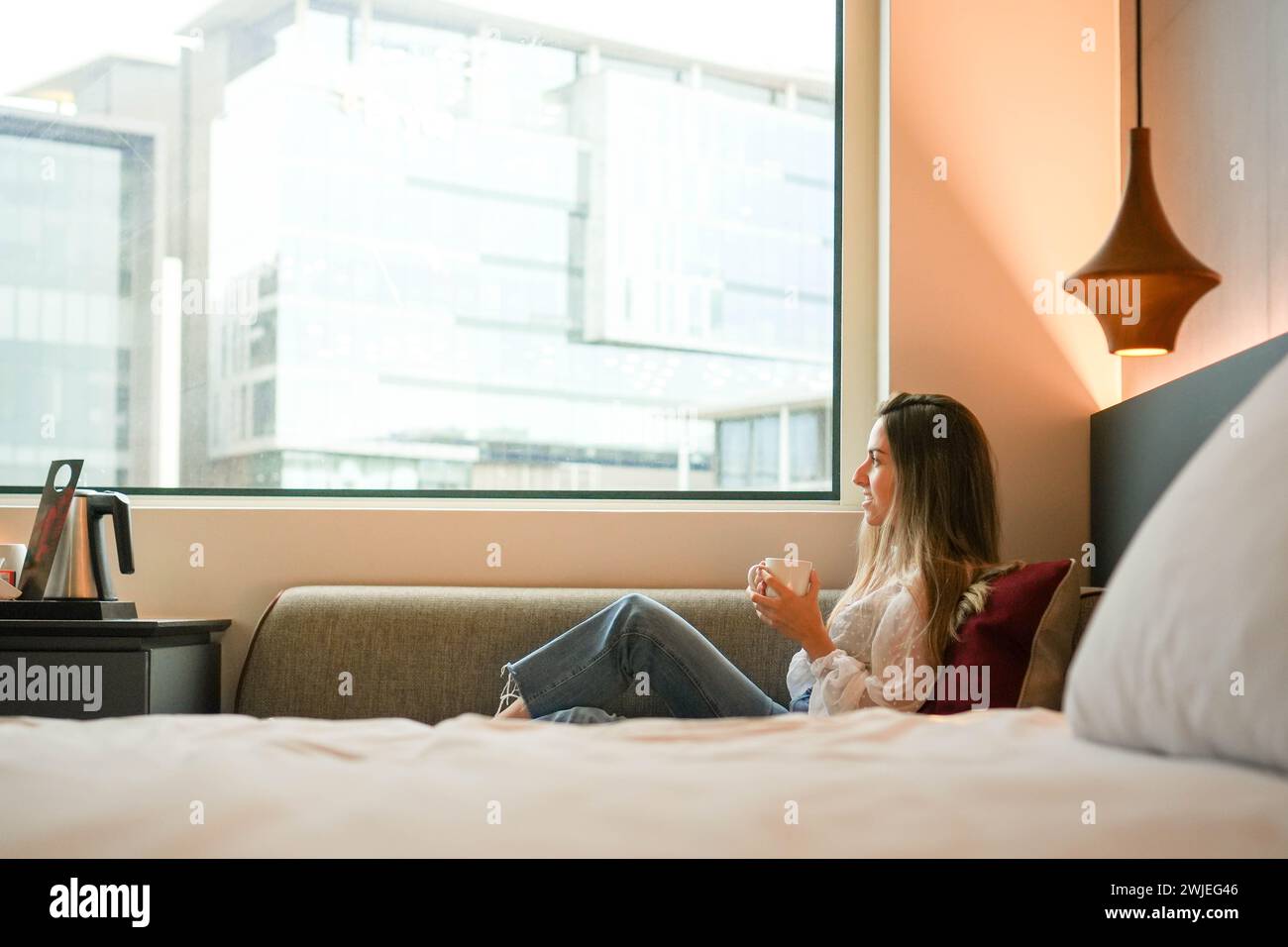 Una giovane donna è seduta comodamente vicino a una finestra in una camera d'albergo, tenendo una tazza con un tranquillo sfondo urbano, suggerendo un momento di relax o di relax Foto Stock