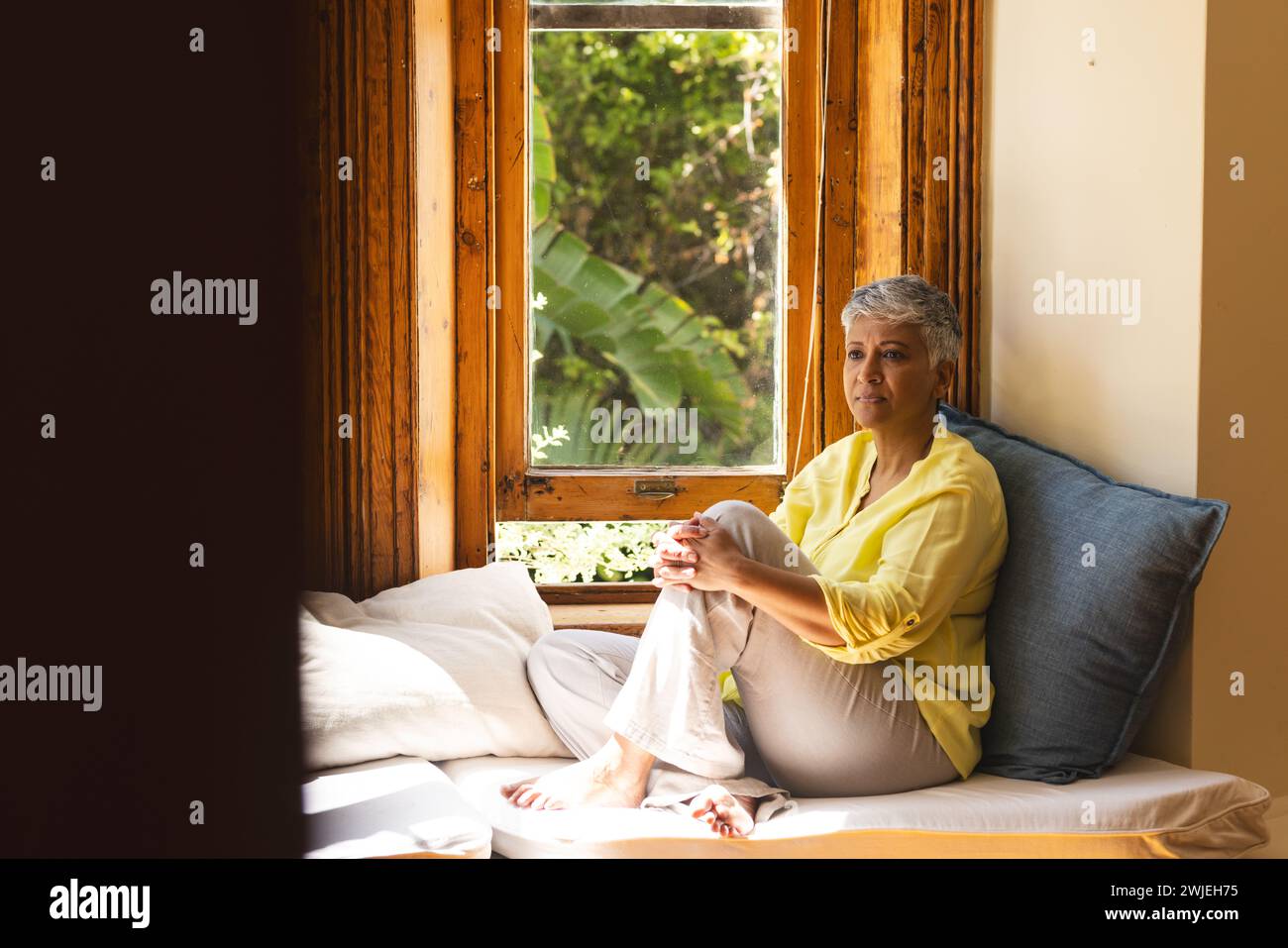 Una donna birazziale matura seduta accanto alla finestra a casa con spazio per le copie Foto Stock