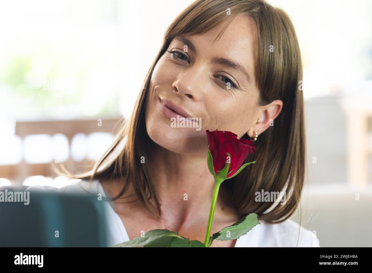 Una giovane donna caucasica sorride dolcemente mentre tiene una rosa rossa vicino al viso Foto Stock
