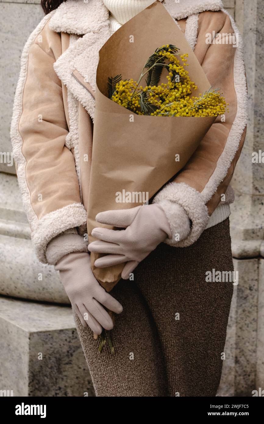 Una donna con una giacca calda beige e guanti in pelliccia che reggono un bouquet di fiori freschi di mimosa. Look invernale elegante Foto Stock