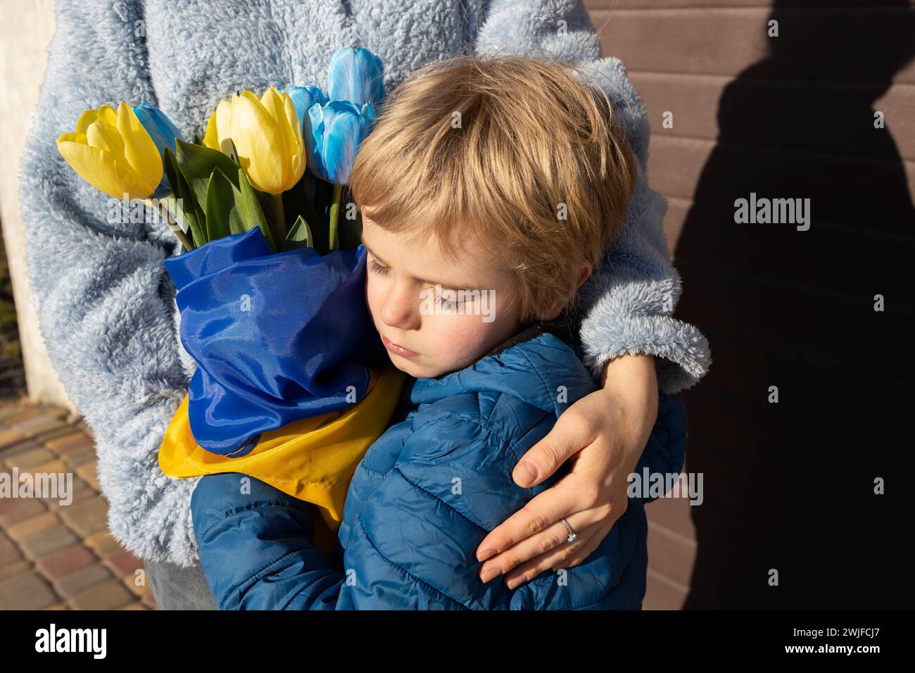 Un bambino triste abbraccia una donna che tiene in mano un mazzo di tulipani gialli e blu avvolti da una bandiera Ucraina. Famiglia, solidarietà, sostegno. Foto Stock