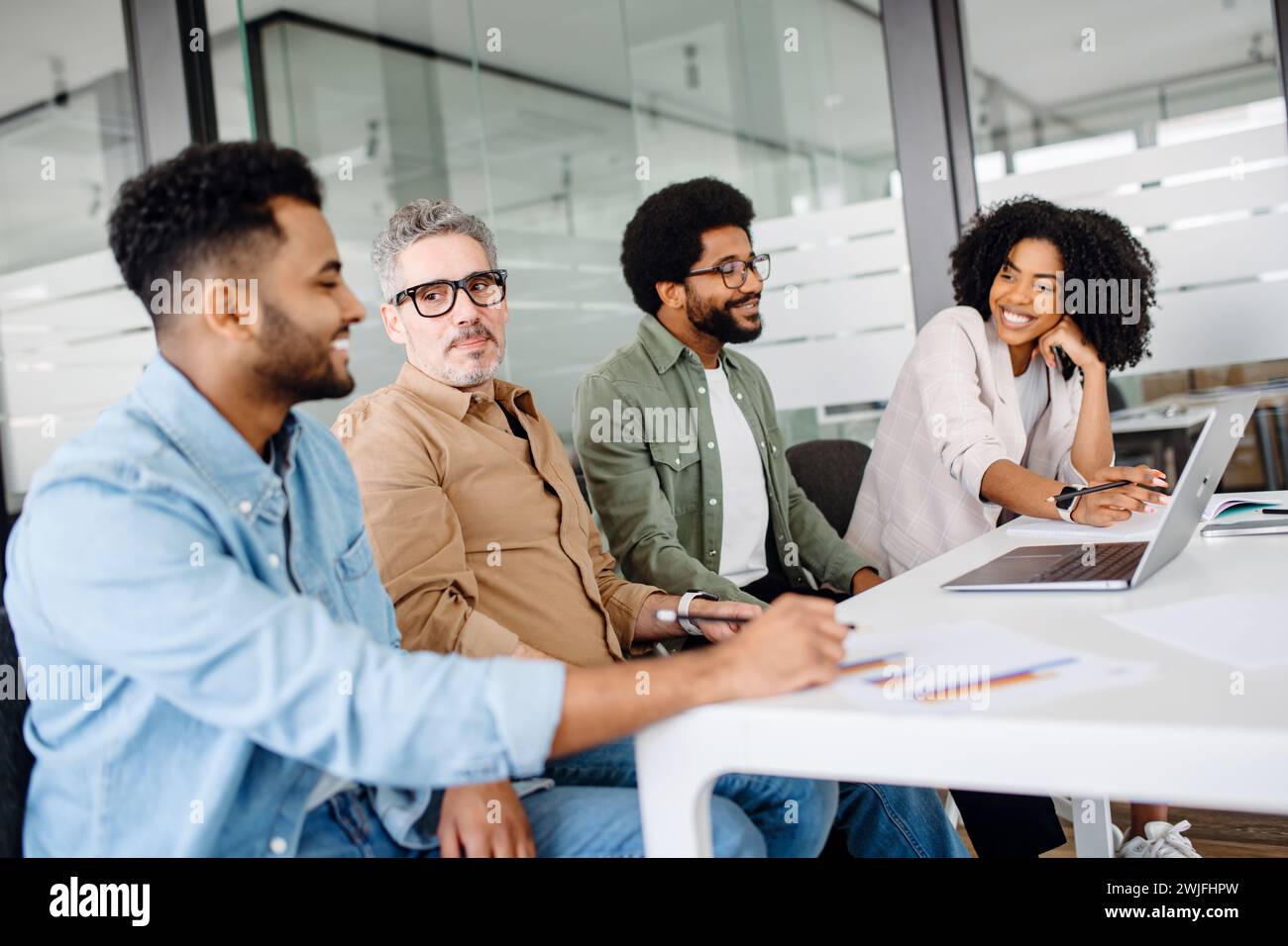 Un gruppo di quattro colleghi si siede casualmente intorno a un tavolo in un moderno ufficio, impegnato in una discussione vivace, con particolare attenzione al lavoro di squadra e alla risoluzione collaborativa dei problemi, sessione di brainstorming Foto Stock