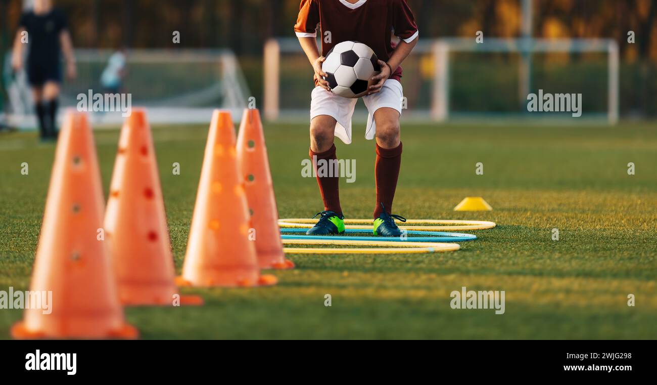 Ragazzo di calcio che tiene la palla in mano e salta sul sentiero degli ostacoli. Sports Agility Hurdles Track. Ragazzo che migliora la velocità e l'agilità Foto Stock