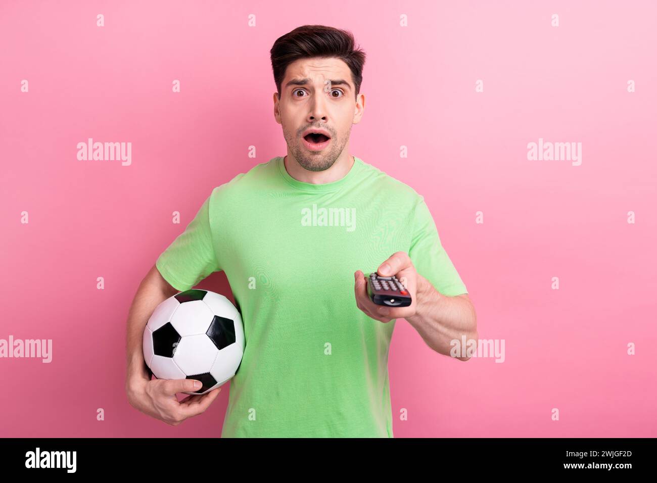 Foto del ragazzo turbato nervoso indossa una maglietta verde tieni la palla da calcio cambiando i canali tv fissando un punteggio negativo isolato su sfondo rosa Foto Stock