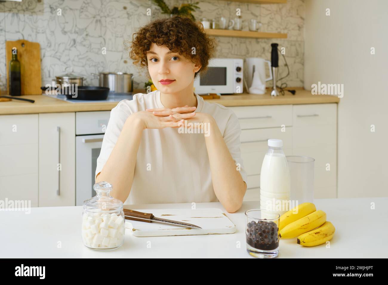 Ritratto di una giovane donna seduta al tavolo della cucina con banane, bottiglia di latte e ribes nero sulla sua superficie Foto Stock