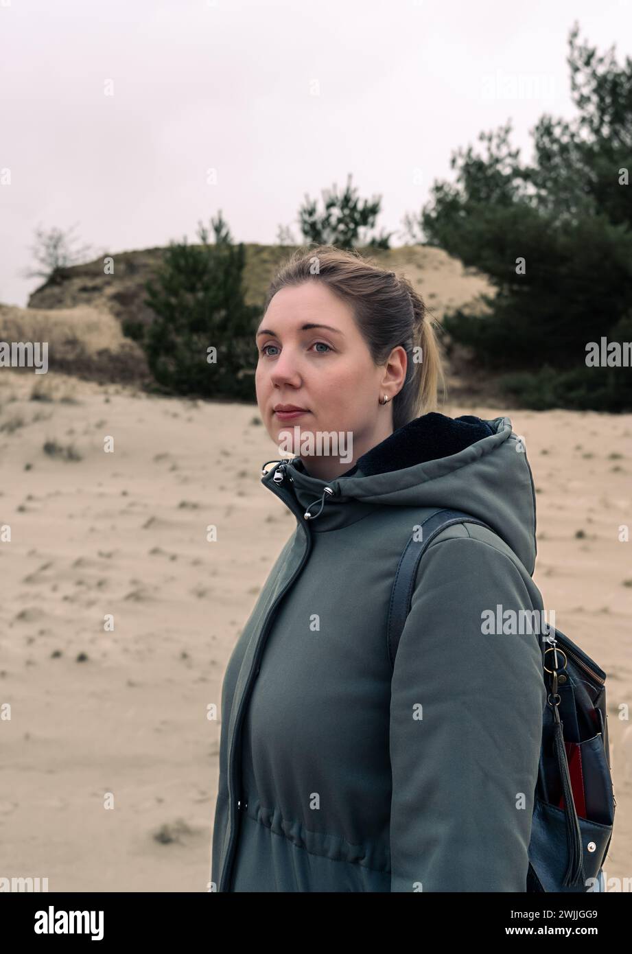 Ritratto di una giovane donna penosa tra le dune di sabbia. I capelli biondi vengono tirati indietro con cura mentre guardano in lontananza con un'espressione premurosa Foto Stock