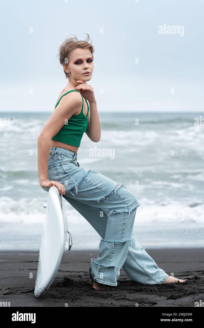 Ritrattista hipster surfista seduta su una tavola da surf distesa sulla sabbia, sullo sfondo delle onde marine. Vista laterale del modello caucasico con capelli corti e trucco Foto Stock