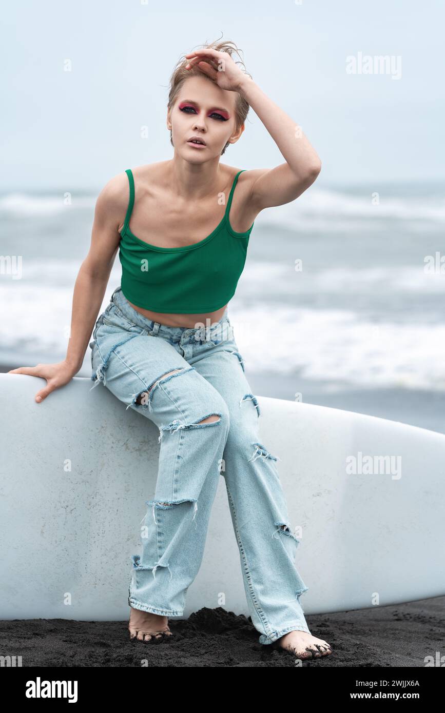 Donna che indossa un top e jeans blu seduti sulla tavola da surf sdraiata sulla sabbia, sullo sfondo delle onde del mare. Ritratto del modello di etnia caucasica hipster Foto Stock