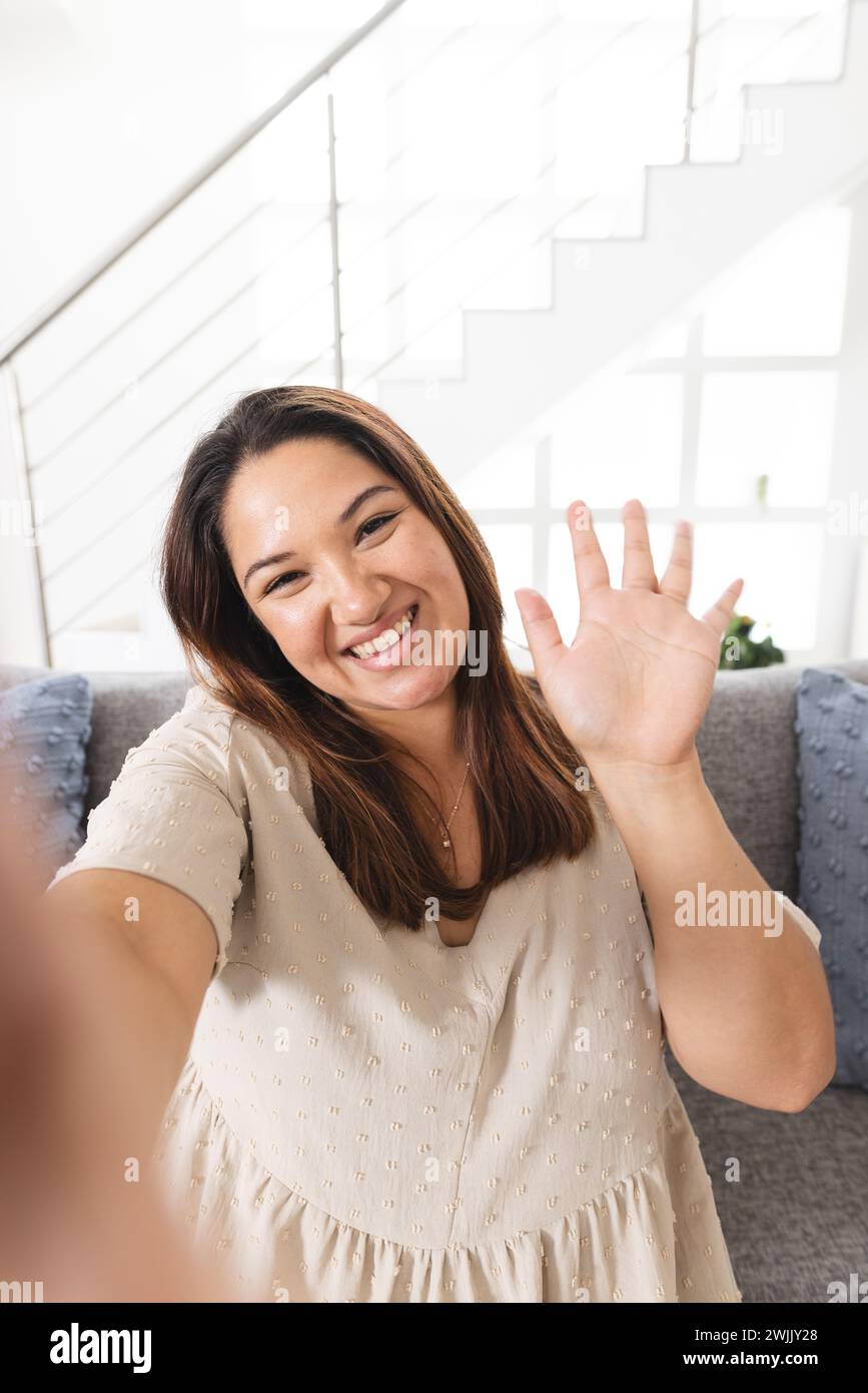 Una giovane donna birazziale ondeggia alla telecamera in un ambiente domestico luminoso durante una videochiamata Foto Stock